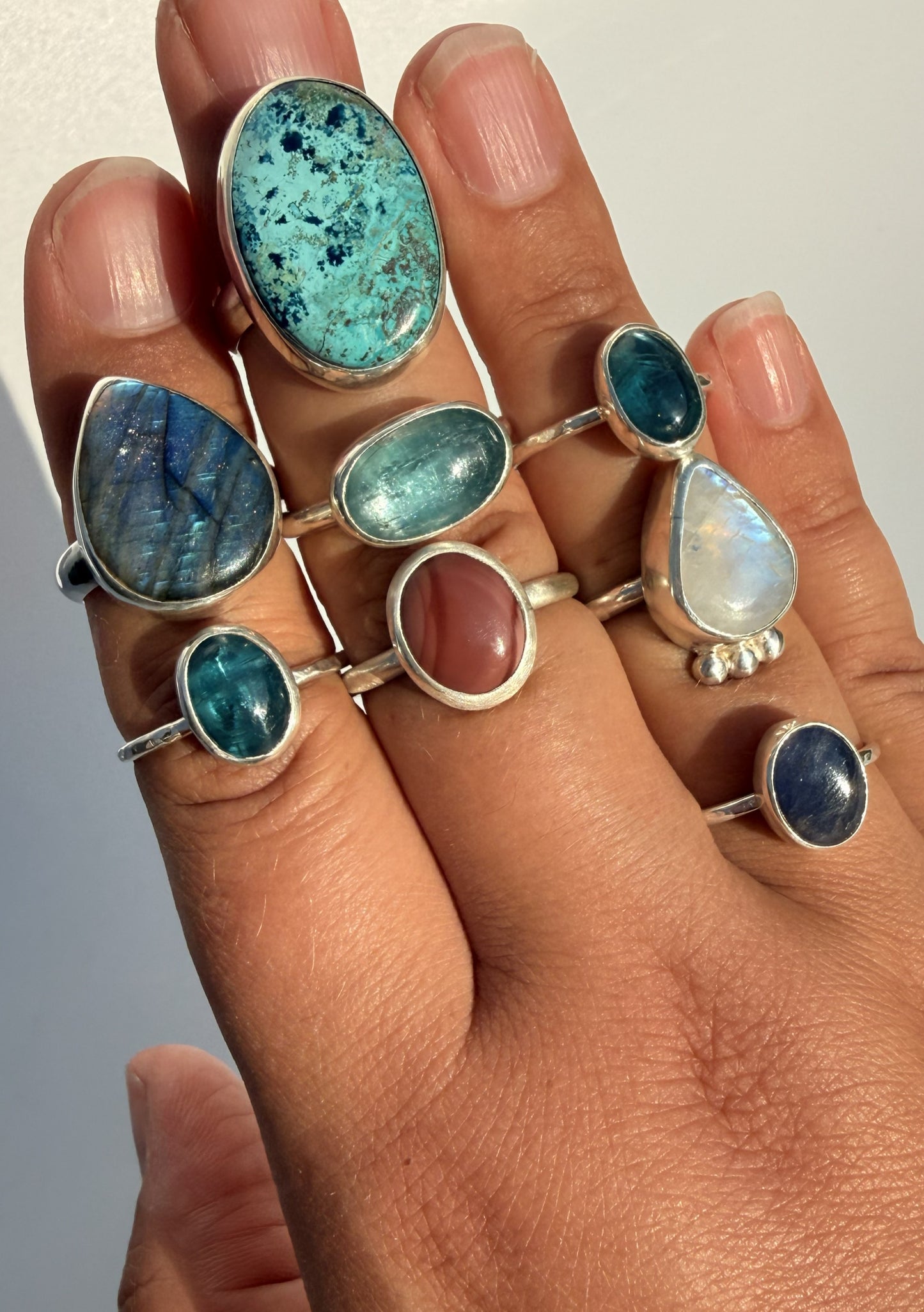 A close-up of a hand wearing multiple silver rings with colorful gemstones, including turquoise, blue, green, pink, and white opal-like stones.