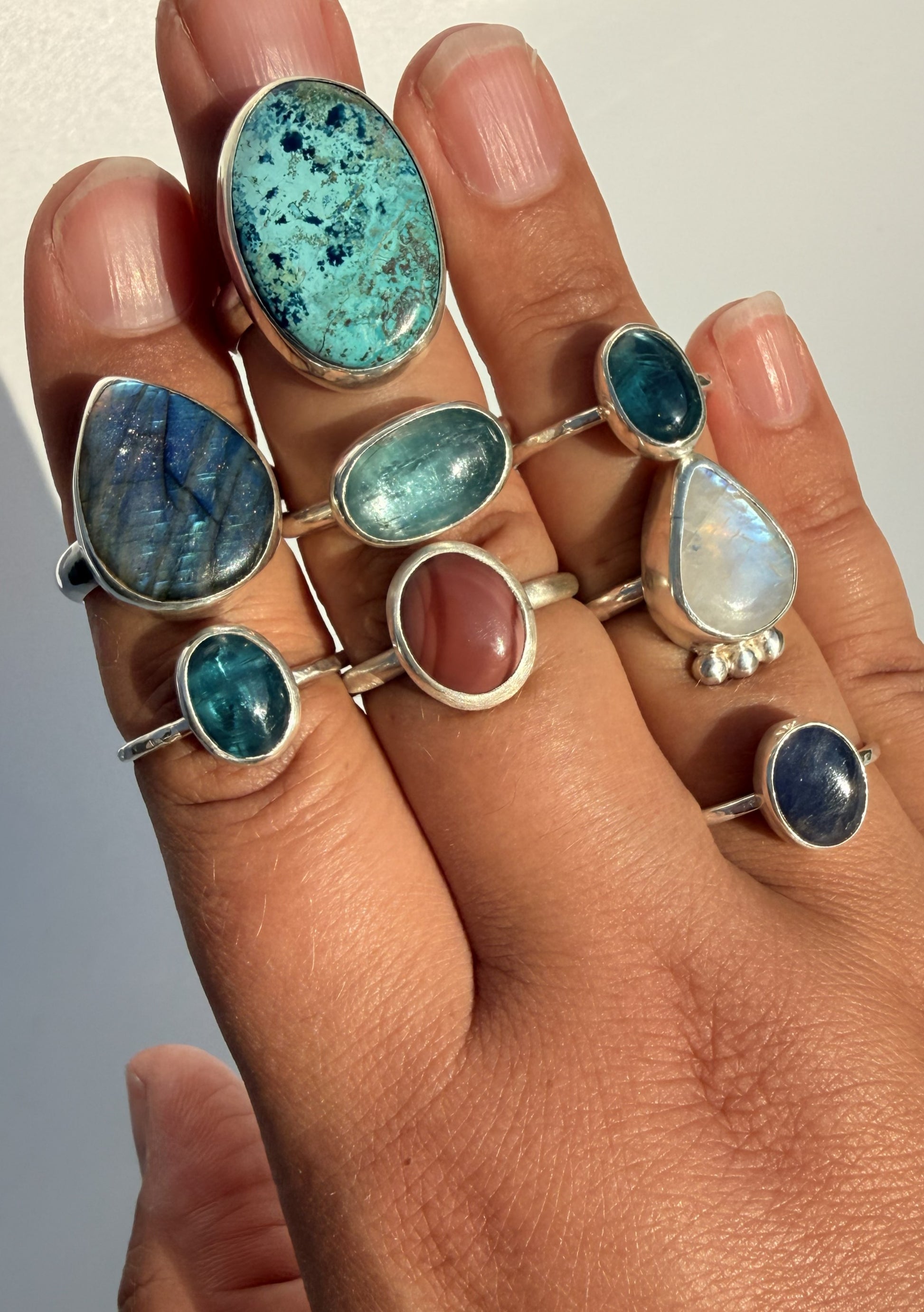 A close-up of a hand wearing multiple silver rings with colorful gemstones, including turquoise, blue, green, pink, and white opal-like stones.