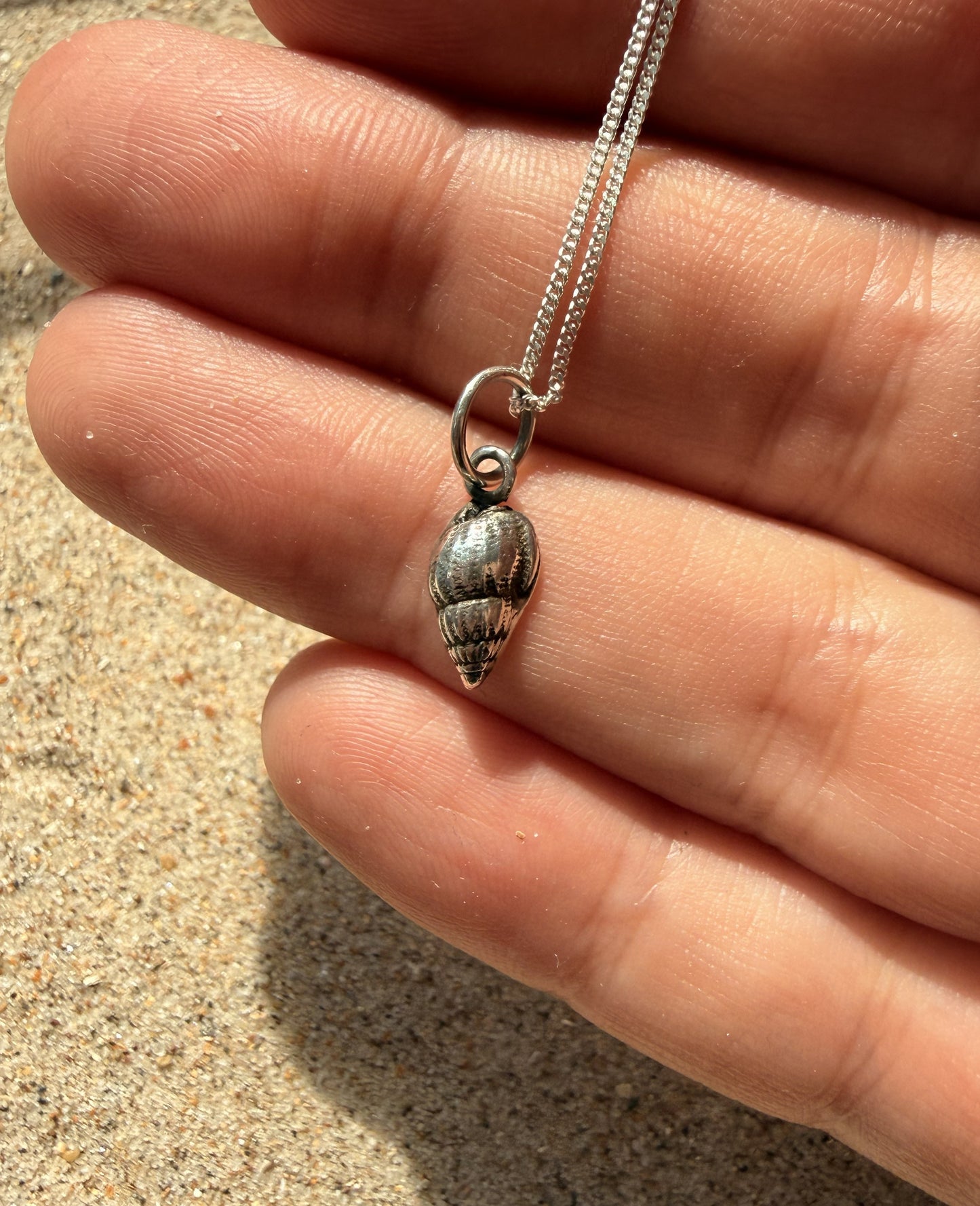 Silver shell pendant on a chain held between fingers with sand in the background