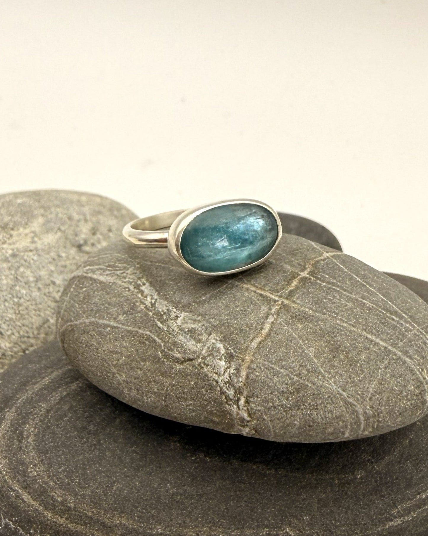 A silver ring with an oval blue-green stone, displayed on a stack of smooth gray river stones against a light beige background.