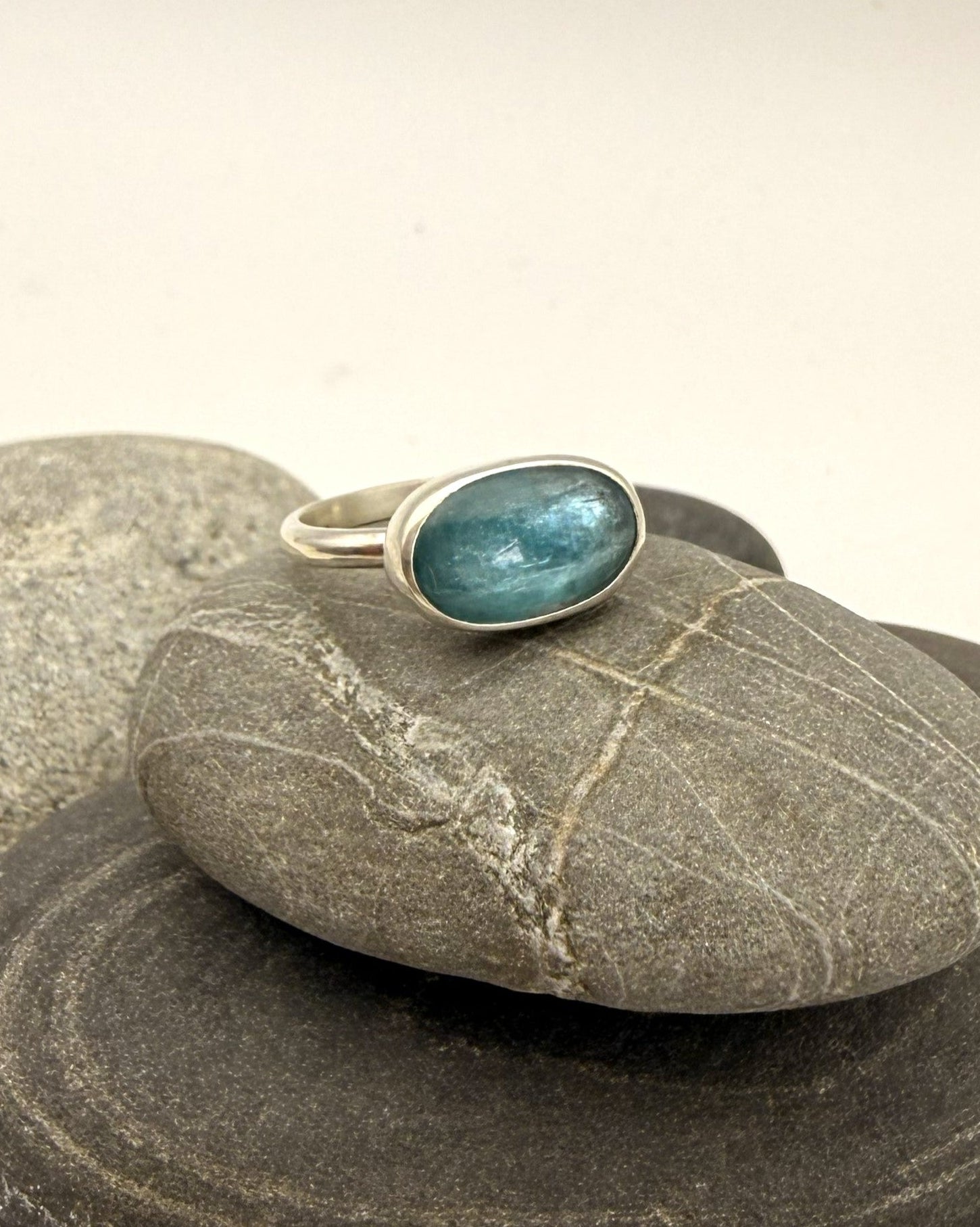 A silver ring with an oval blue-green stone, displayed on a stack of smooth gray river stones against a light beige background.