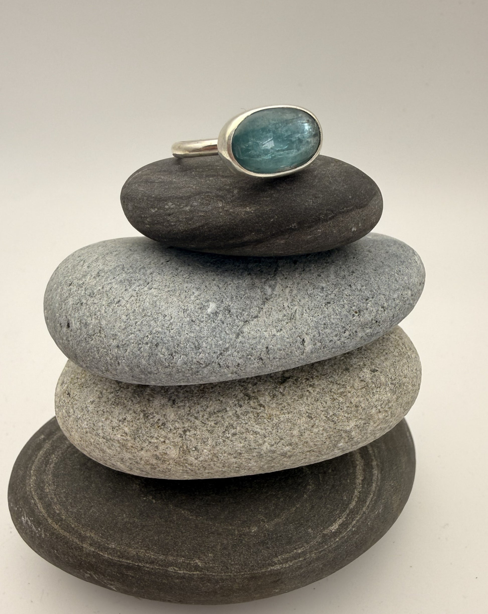 A silver ring with a round teal-blue gemstone is displayed on a stack of four smooth gray and dark gray stones against a light gray background.