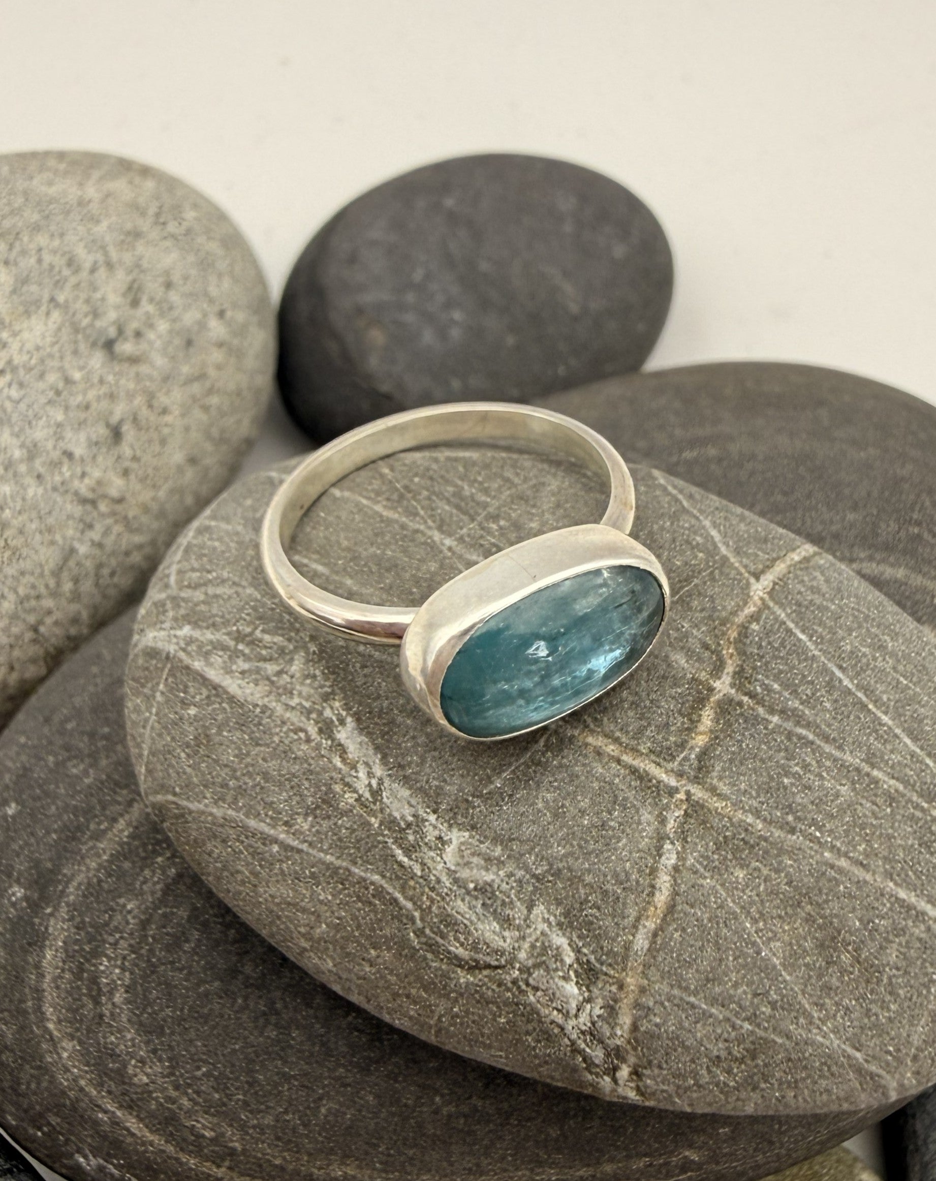 A silver ring with a bezel-set oval blue-green stone, displayed on smooth gray decorative stones against a light neutral background.