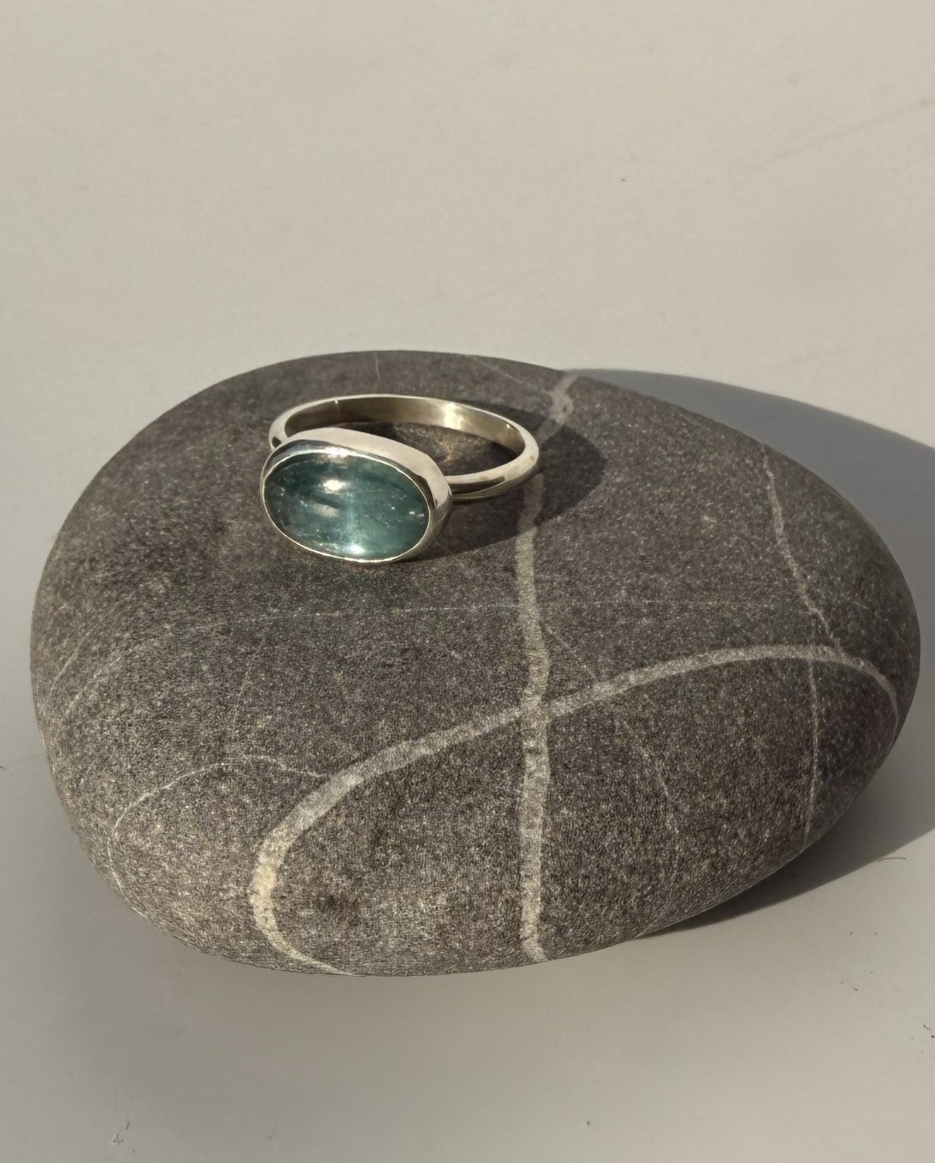 A silver ring with an oval green gemstone, displayed on a smooth gray stone against a neutral light gray background.