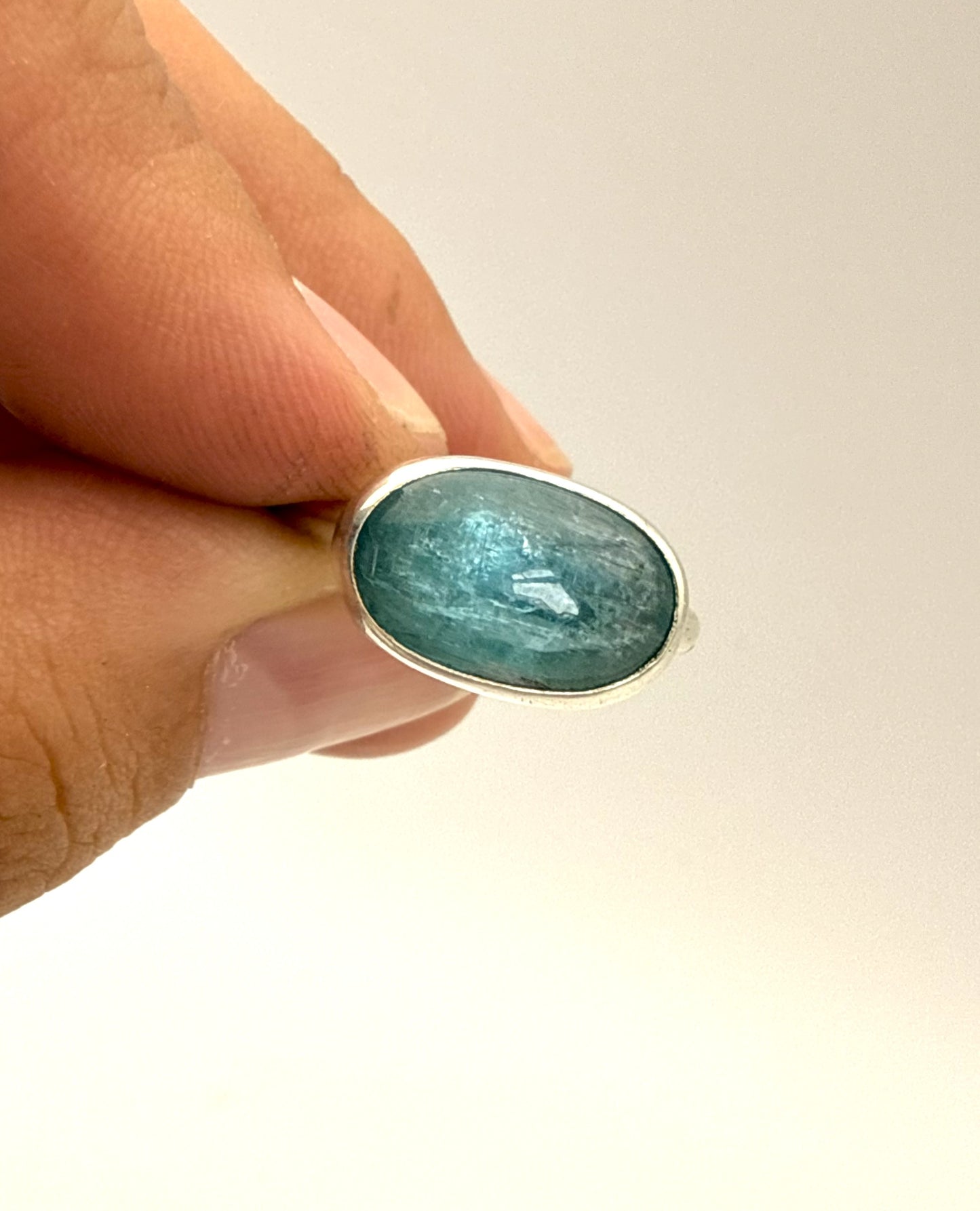 A close-up of a hand holding a silver ring with an oval-shaped light blue gemstone against a plain light background.
