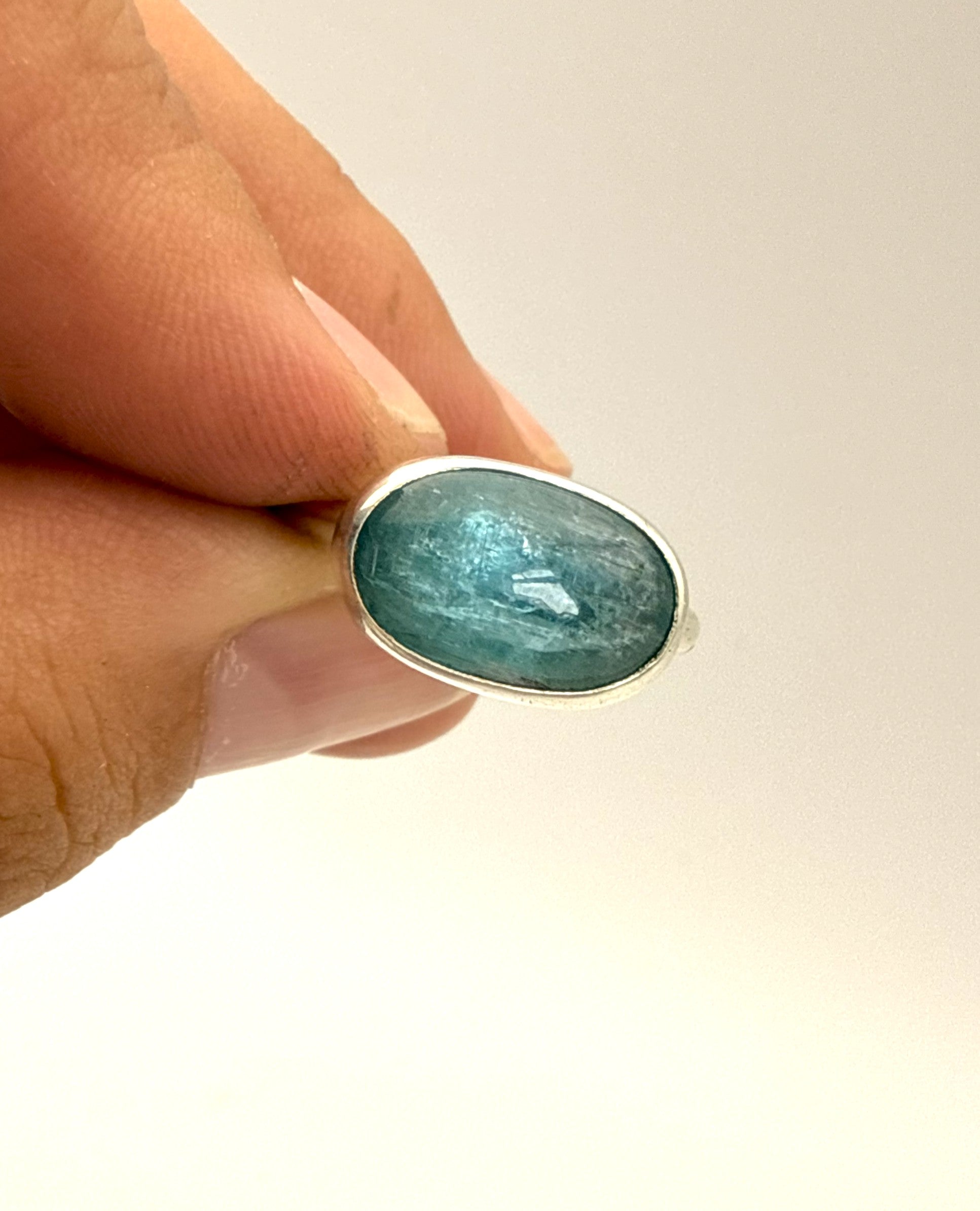 A close-up of a hand holding a silver ring with an oval-shaped light blue gemstone against a plain light background.