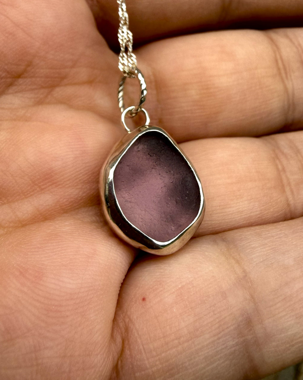 A close-up of a hand holding a silver pendant necklace with an irregular, organic-shaped purple glass stone on a silver chain.