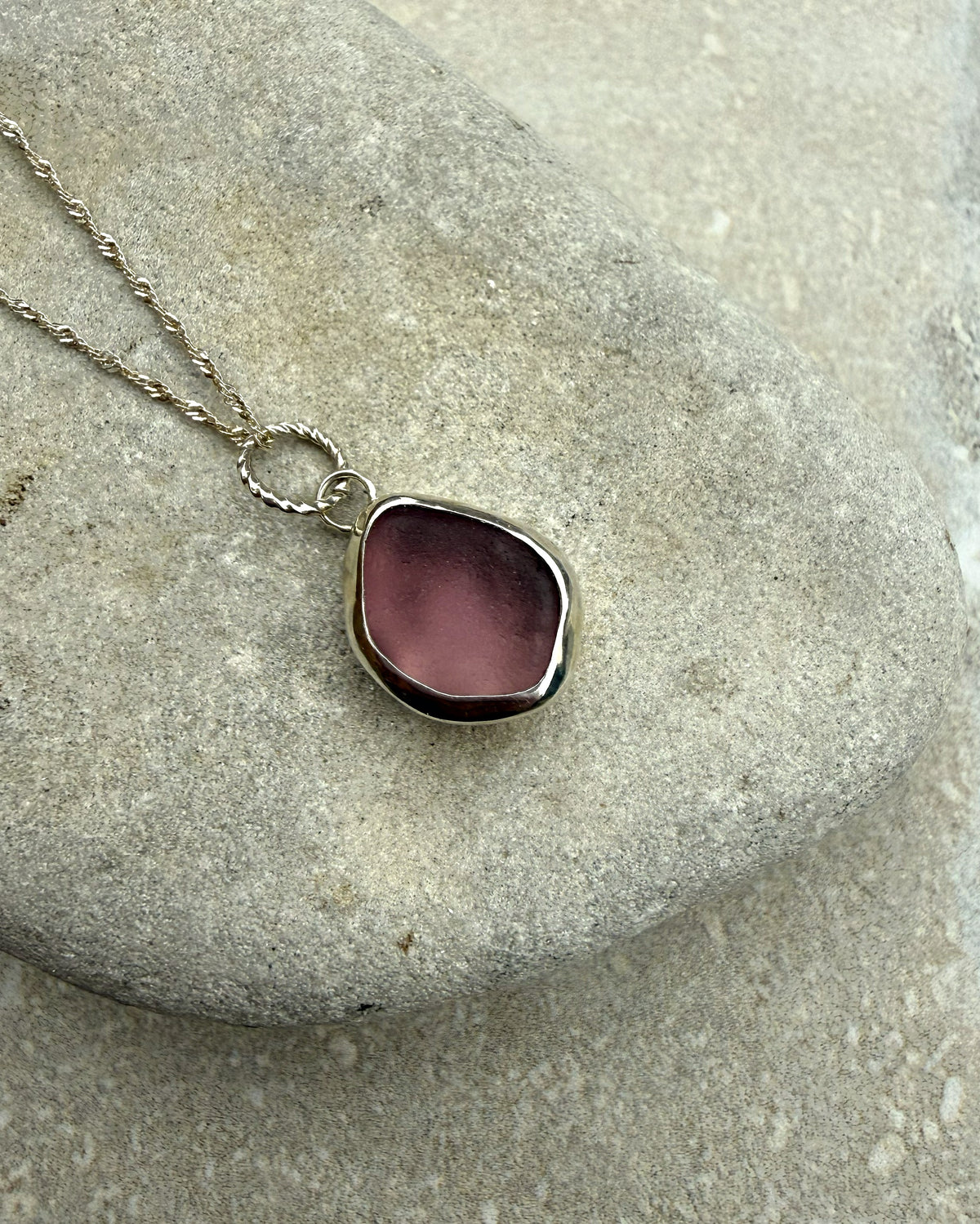 A silver-toned necklace with a polished, irregular-shaped dusty pink pendant resting on a smooth gray stone.