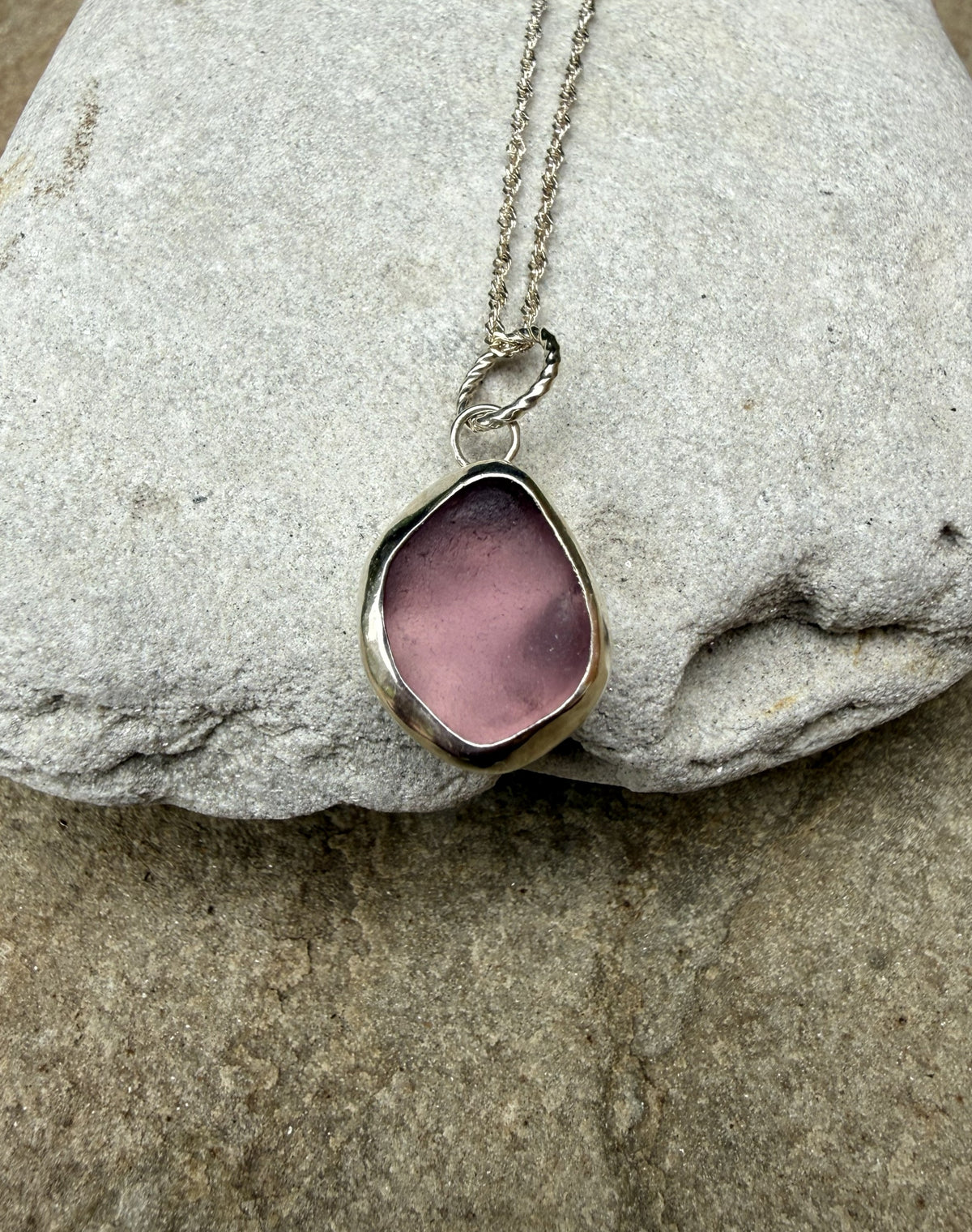 A silver-toned necklace with an irregular, organic-shaped pink glass or stone pendant, displayed on a textured gray stone surface.