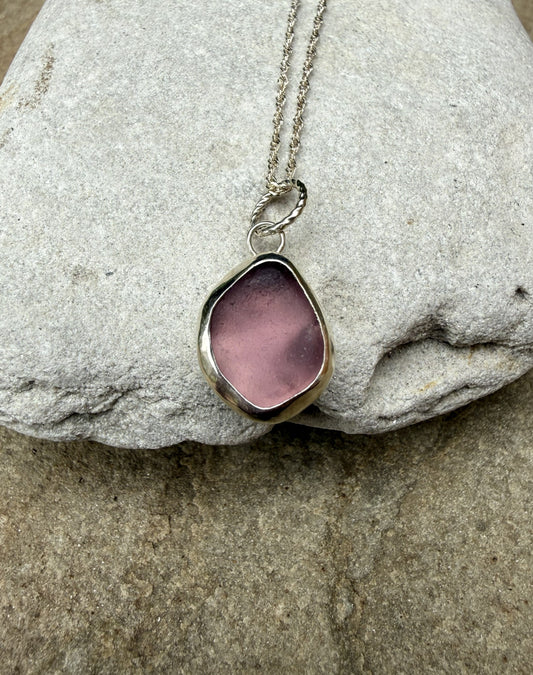A silver-toned necklace with an irregular, organic-shaped pink glass or stone pendant, displayed on a textured gray stone surface.