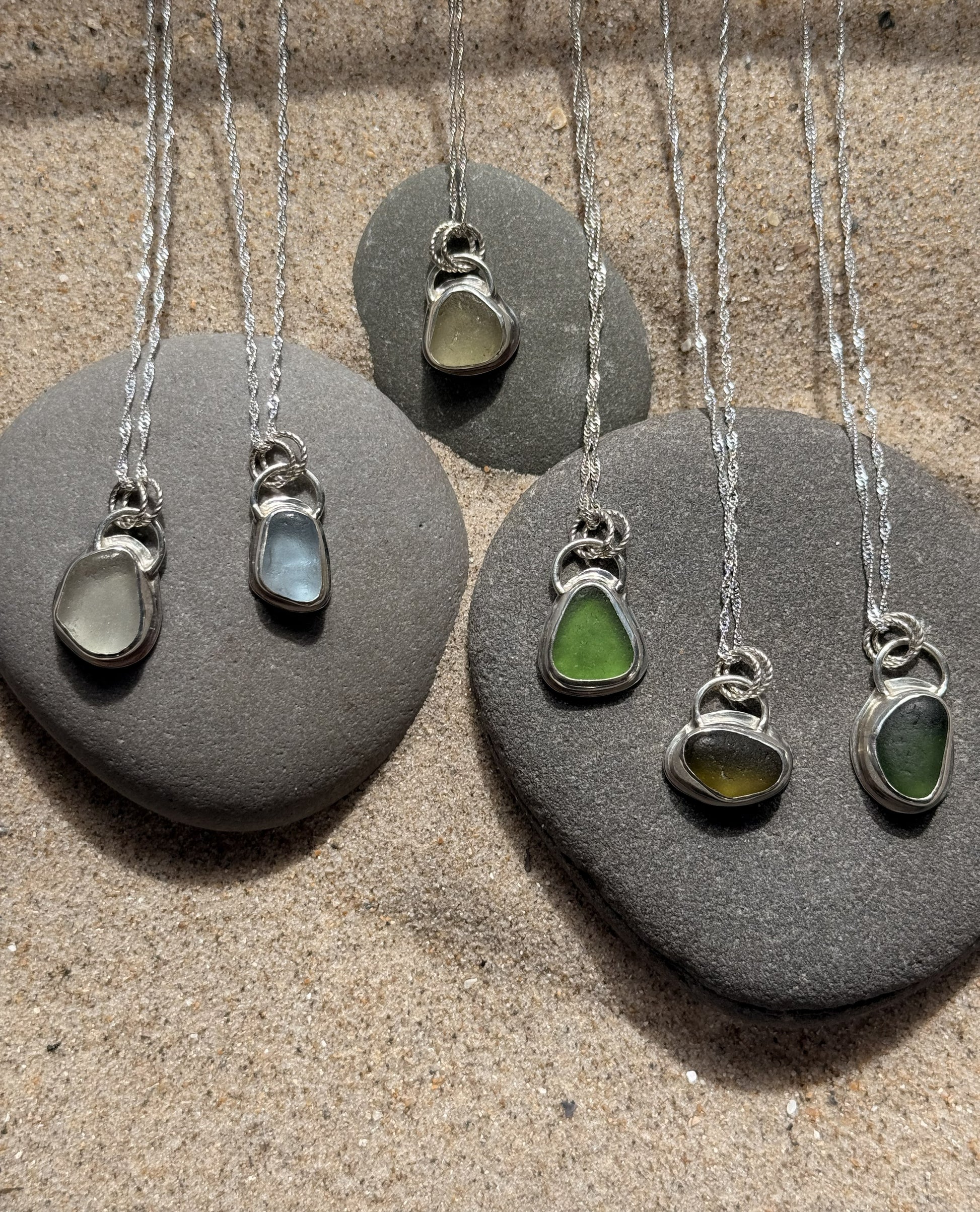 Five sea glass pendants on silver necklaces displayed on gray stones.