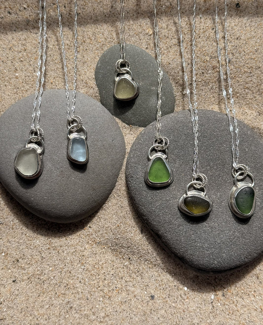 Five sea glass pendants on silver necklaces displayed on gray stones.