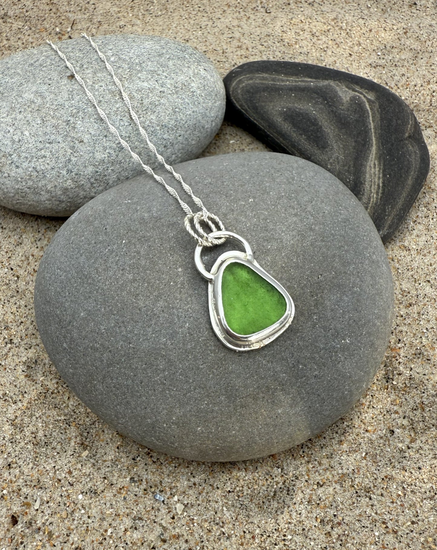 Green sea glass necklace on a gray stone with sand and pebbles in the background