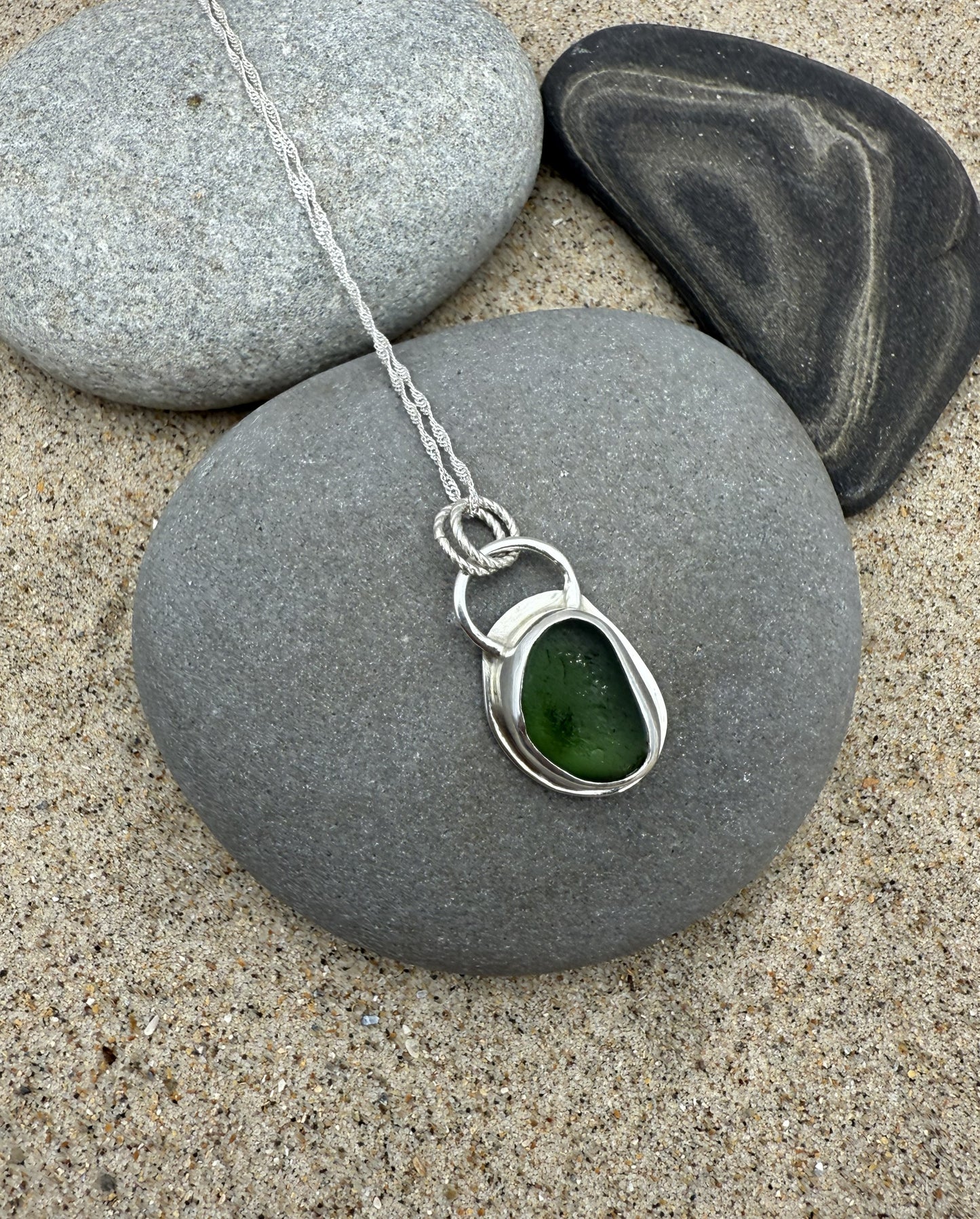 Green teardrop pendant on a silver chain on a stone with sand and pebbles in the background