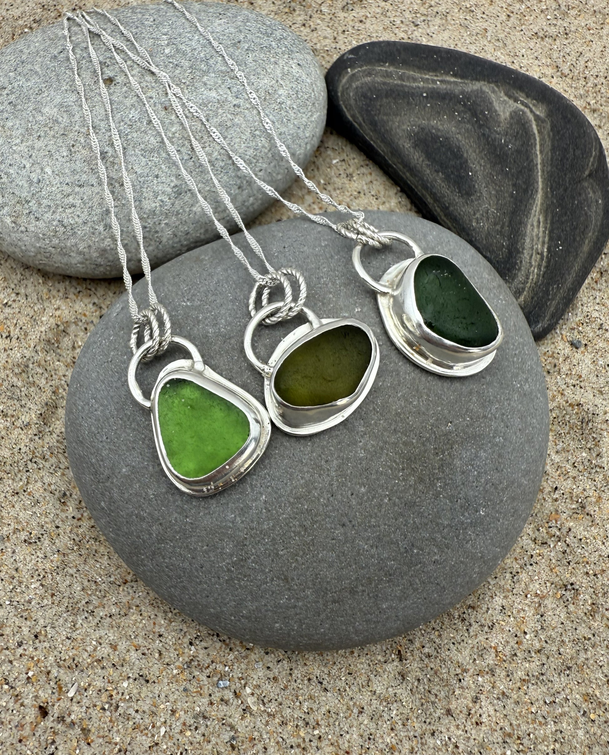 Green sea glass pendants in silver settings on a stone with sand background