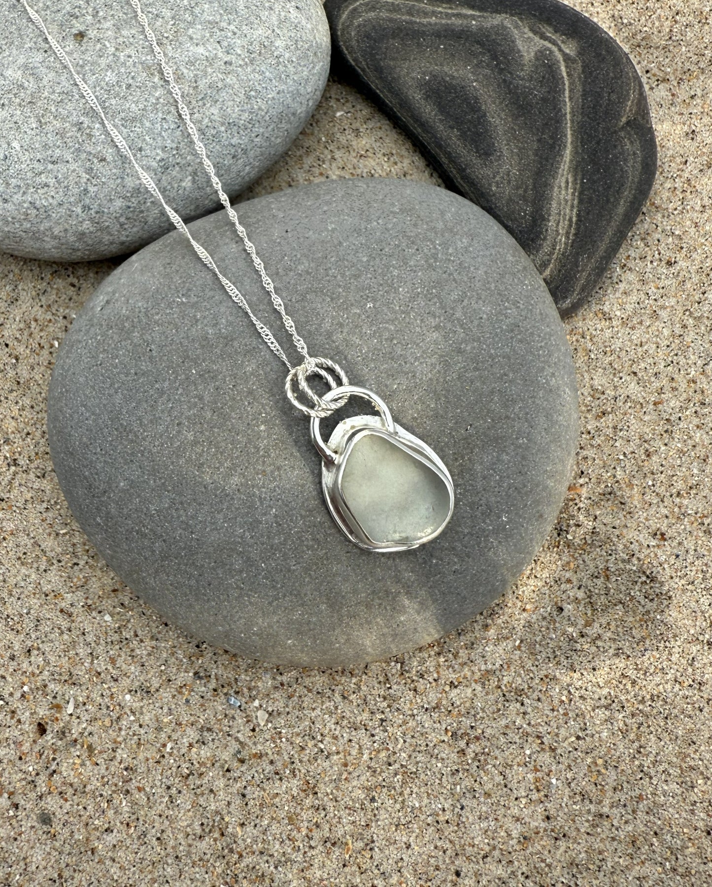 Necklace with a silver sea glass pendant on a stone with sand and pebbles in the background