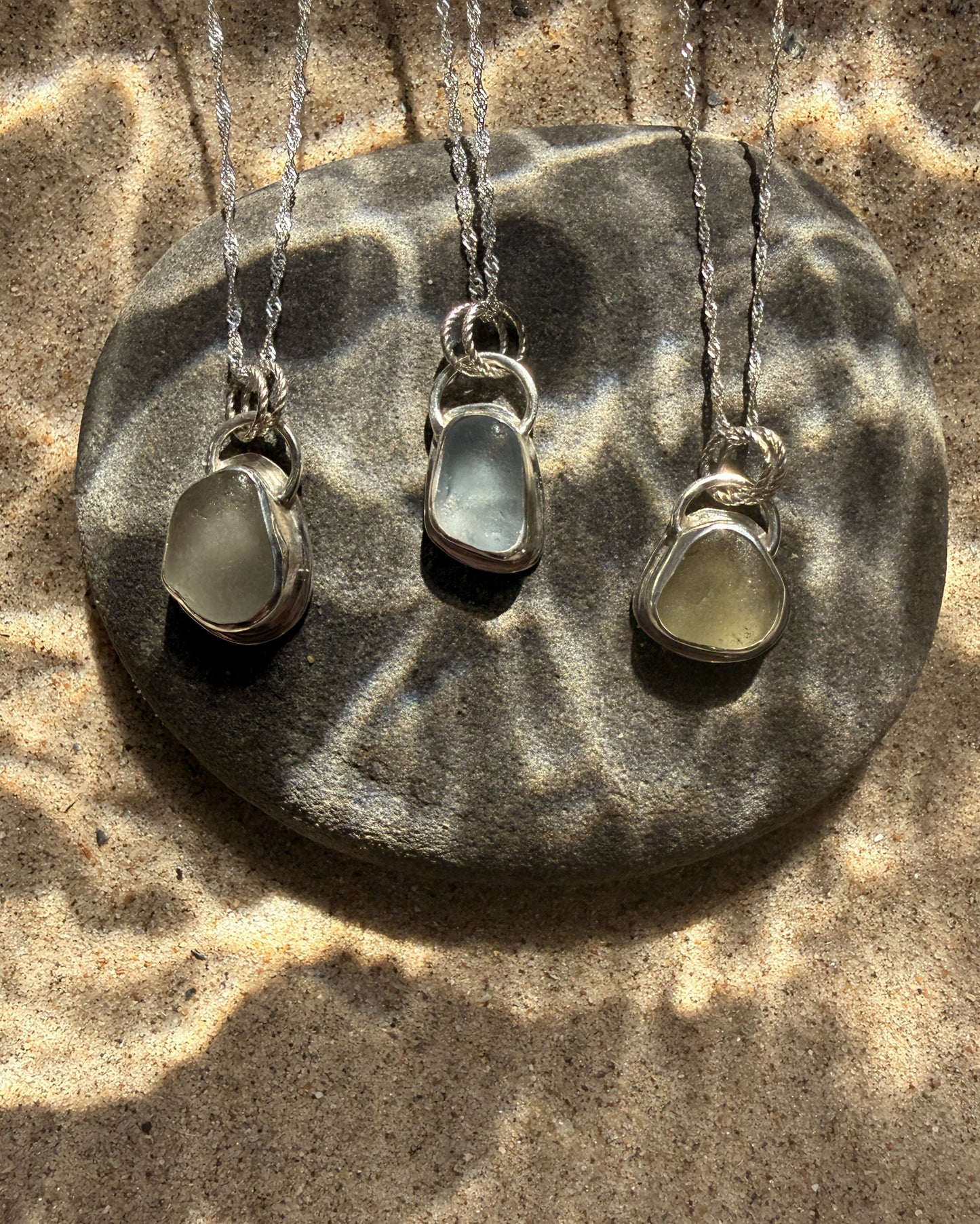 Three sea glass pendants on silver necklaces displayed on a stone surface.