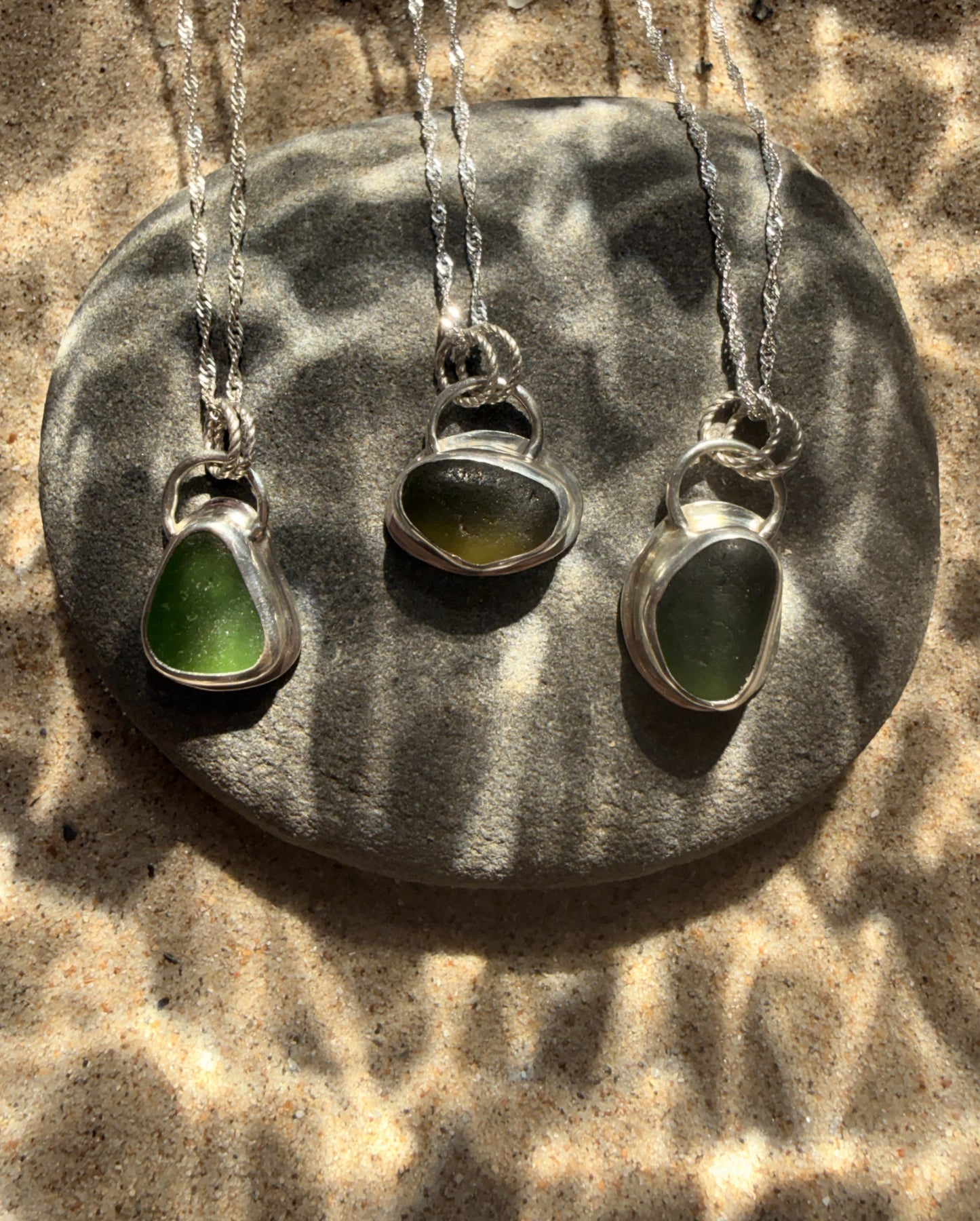 Three silver necklaces with green pendant stones on a textured stone surface.