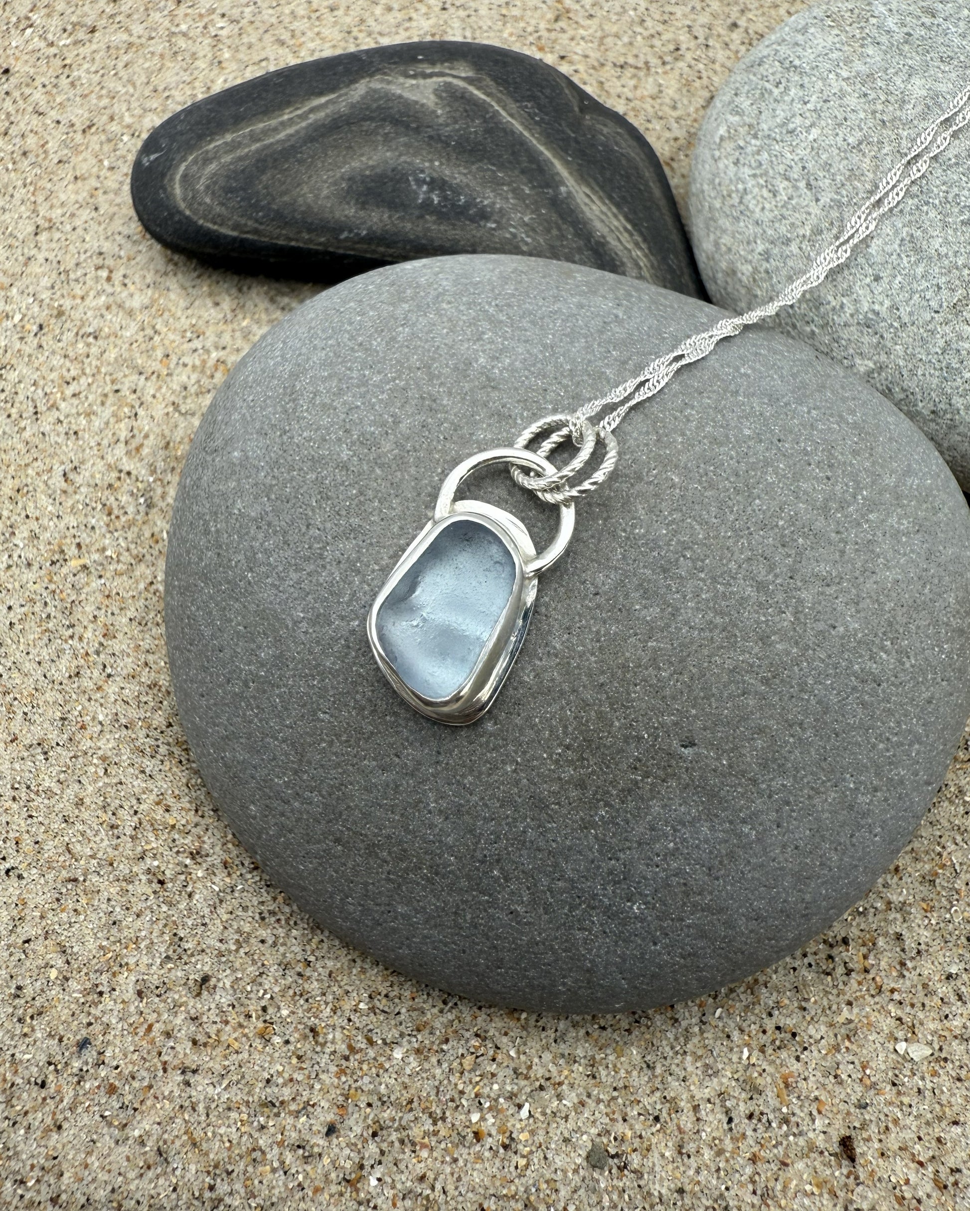 Silver sea glass necklace with a pendant on a stone with sand and pebbles in the background