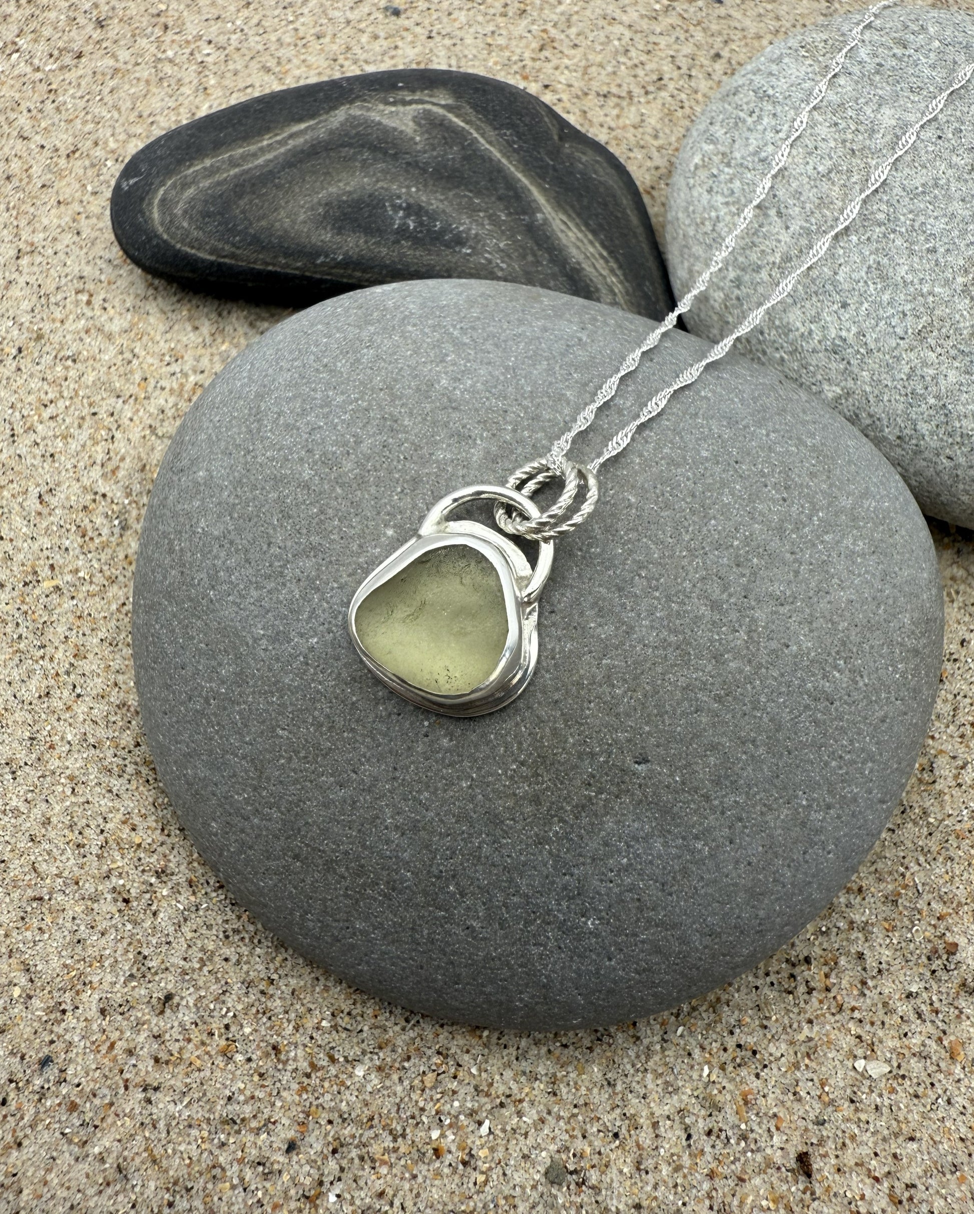 Necklace with a green sea glass pendant on a gray stone with sand and rocks in the background
