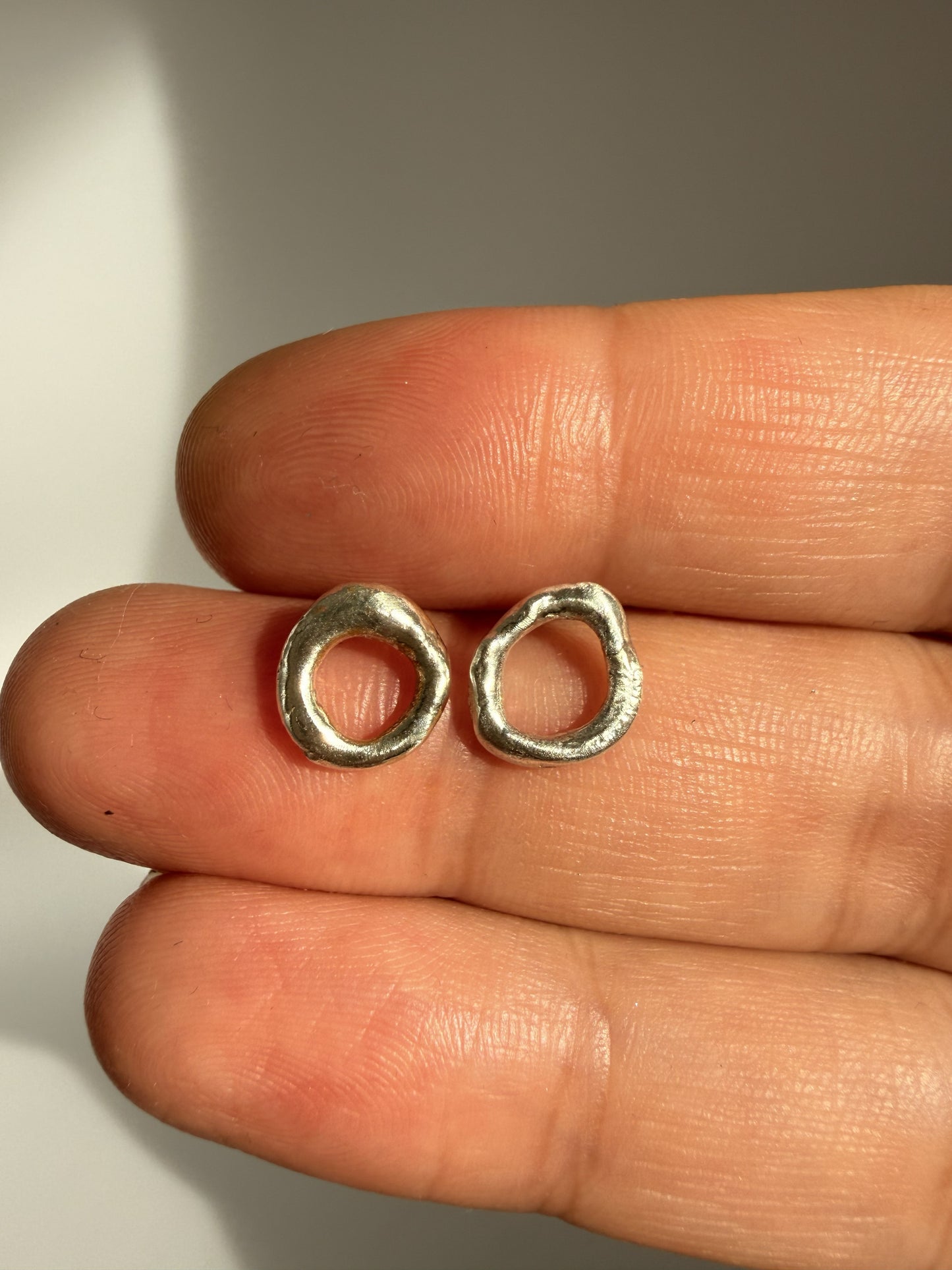 Two small silver molten silver earrings held between fingers on a neutral background