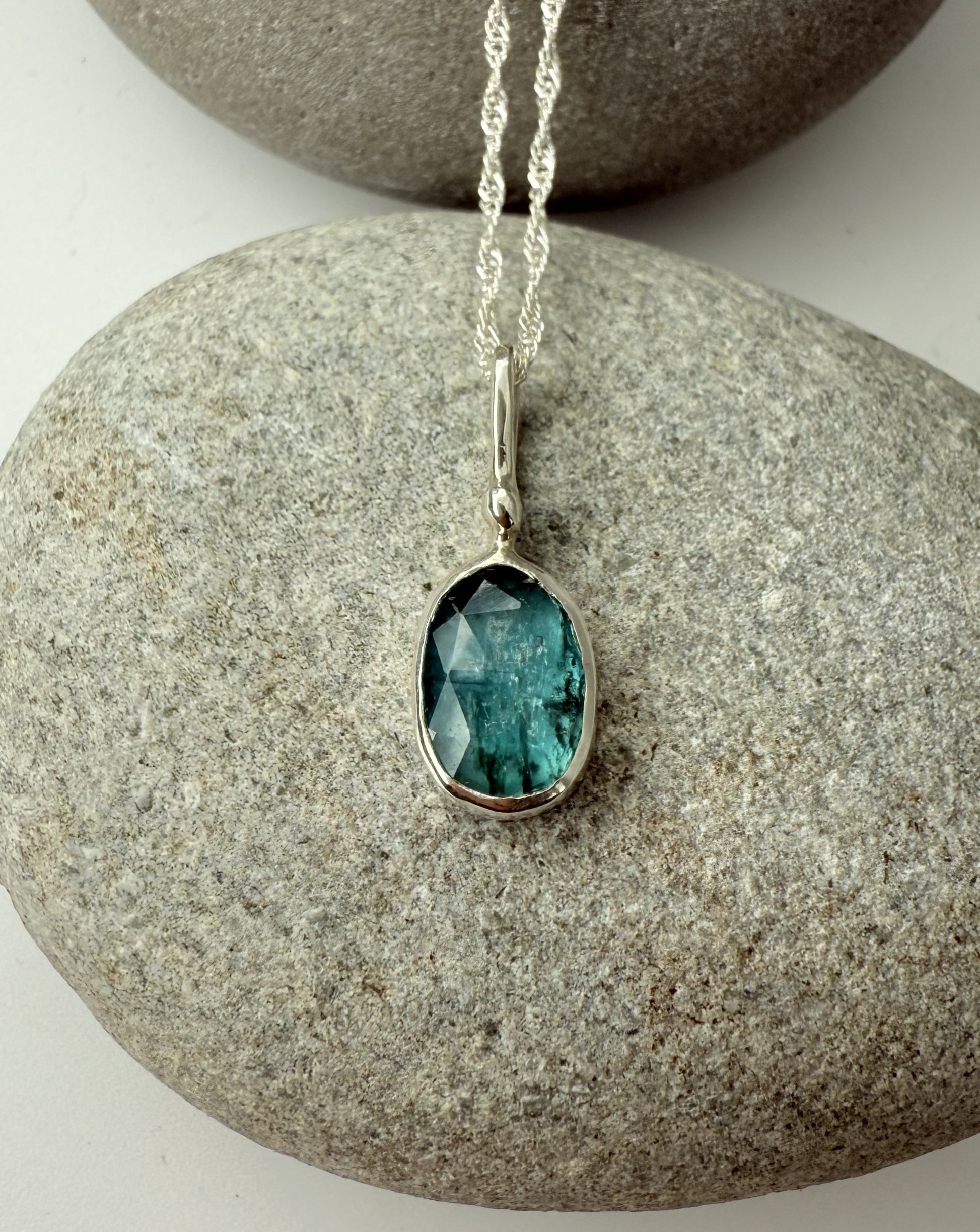 Teal kyanite gemstone pendant on a silver chain resting on a stone surface