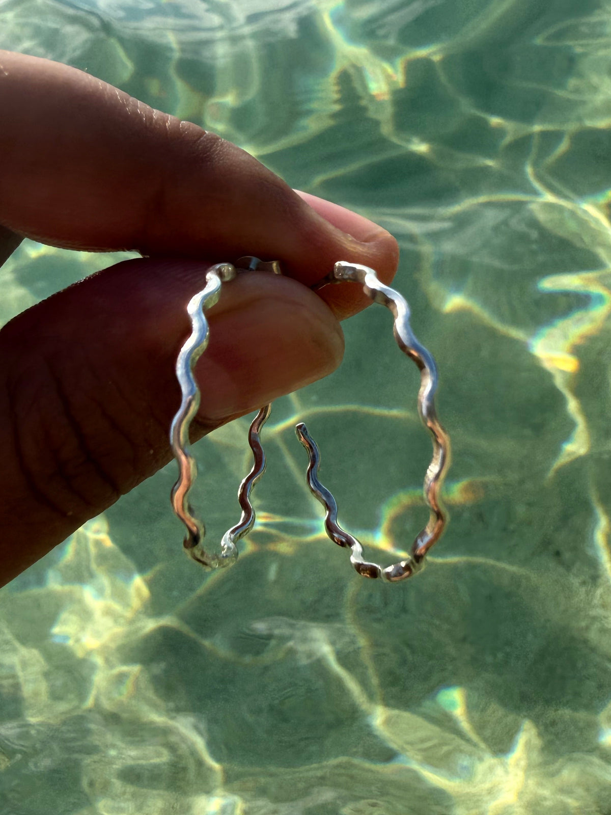 A hand holding a pair of silver wavy hoop earrings against a background of clear, sun-dappled ocean water.
