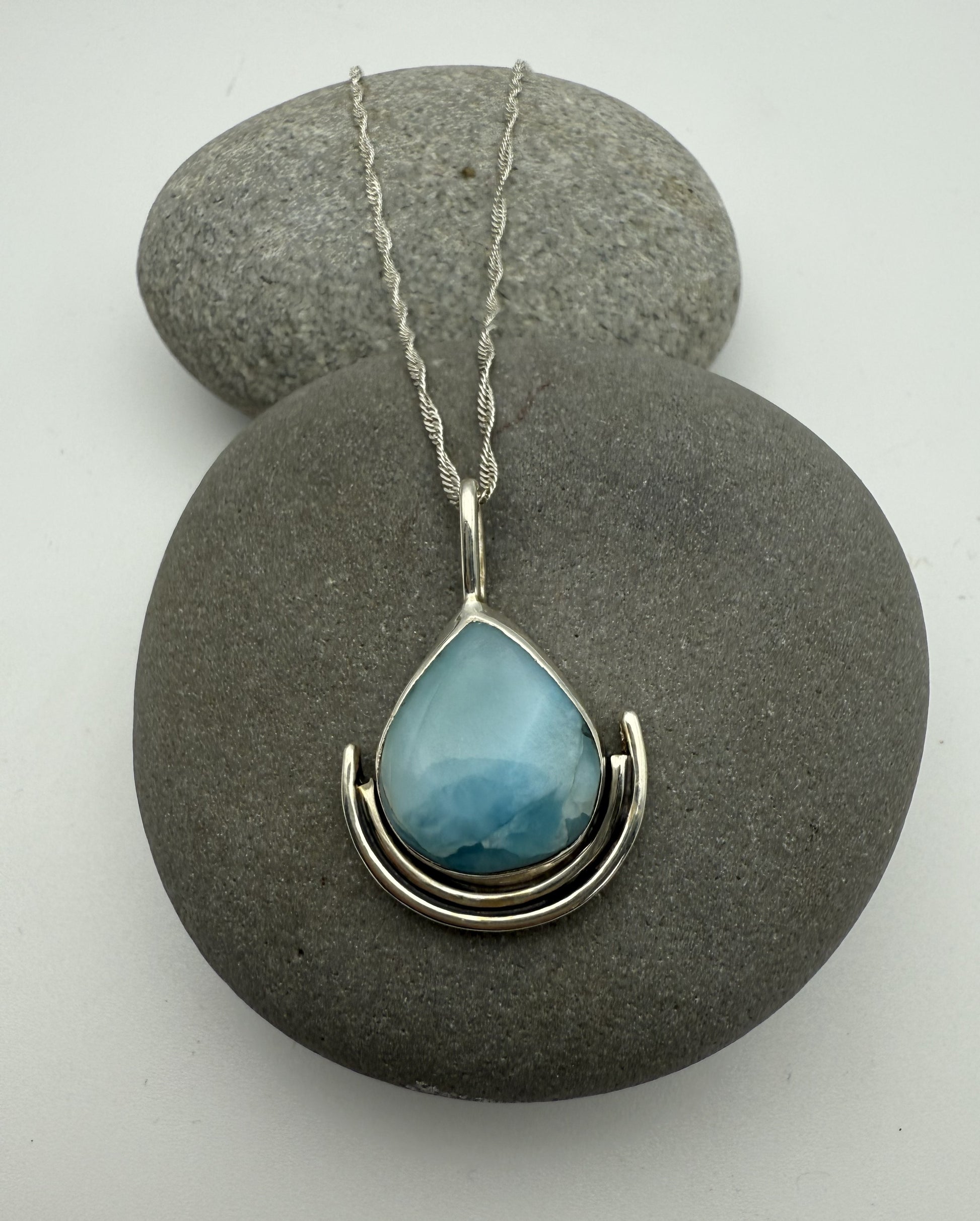 Teardrop-shaped blue larimar pendant on a silver chain resting on grey stones.