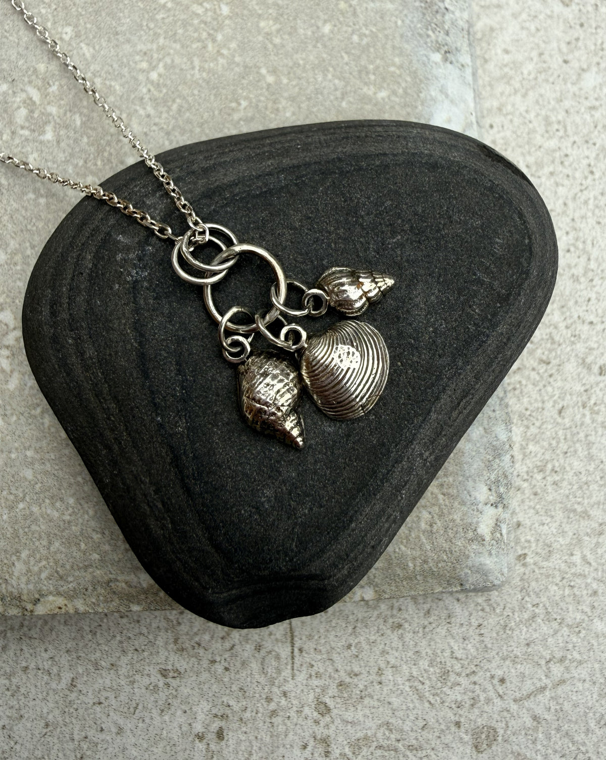 A silver-toned necklace with three seashell charms resting on a dark, polished black stone against a light gray textured background.