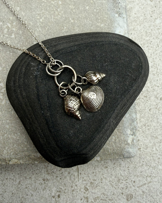A silver-toned necklace with three seashell charms resting on a dark, polished black stone against a light gray textured background.