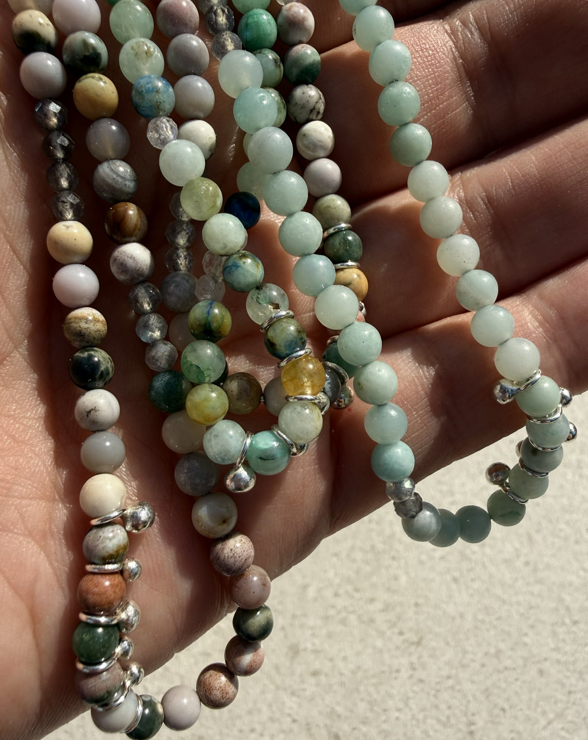 A close-up of a hand holding several strands of polished gemstone beads, including light green, white, brown, gray, and marbled varieties, with silver spacer beads.