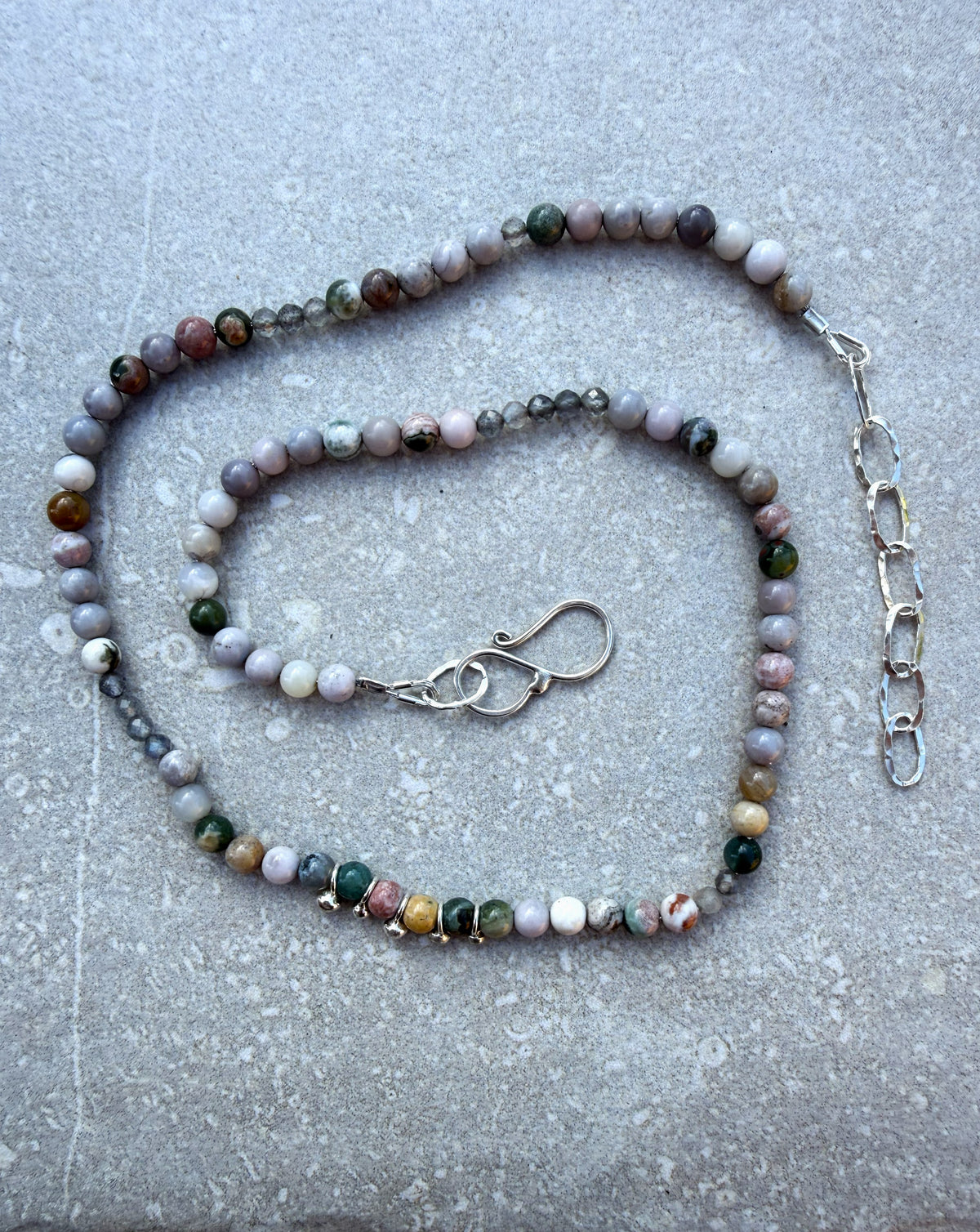 A beaded necklace made of polished round gemstone beads in earthy, marbled colours, displayed on a textured grey surface. The beads feature shades of grey, pink, green, brown, and white.