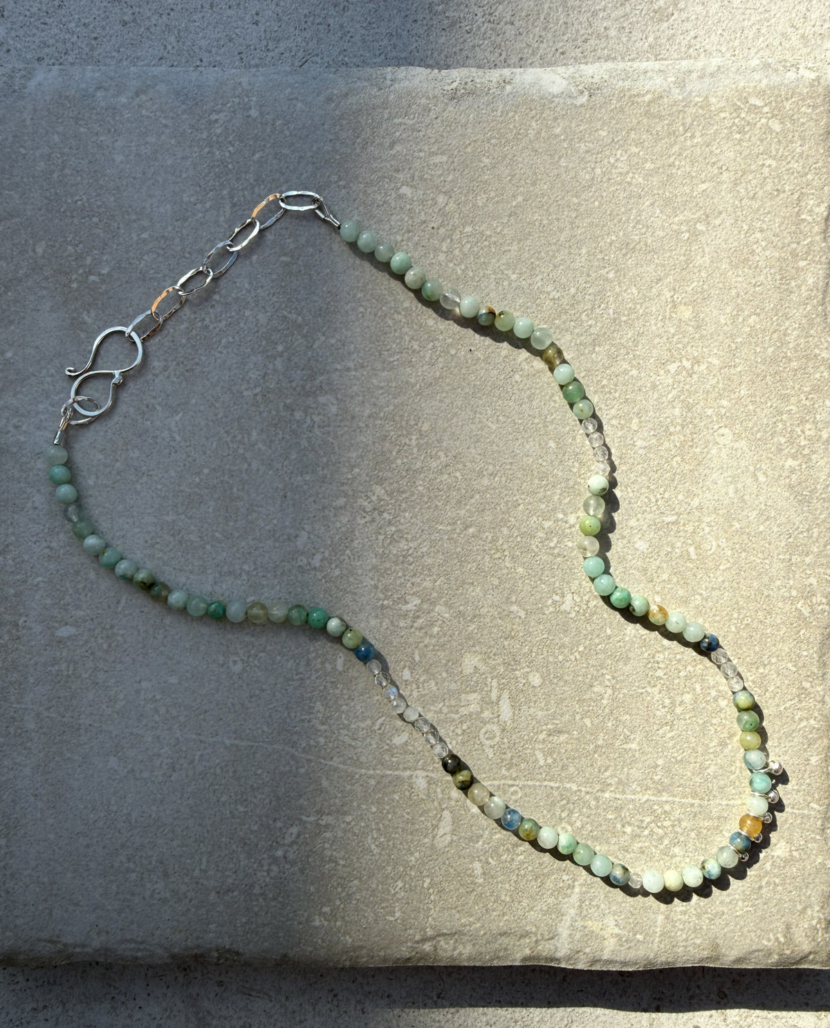 A beaded necklace made of small green, blue, and white marbled beads with a silver lobster clasp, laid out on a textured gray stone surface.