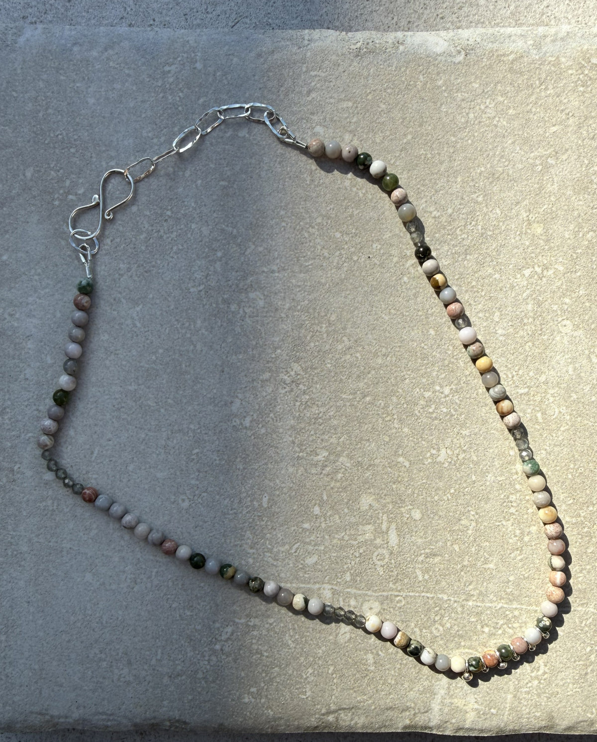 A beaded necklace made of small round beads in muted pink, grey, green, and brown tones, with a silver-toned chain extender and lobster clasp, laid on a textured stone surface.