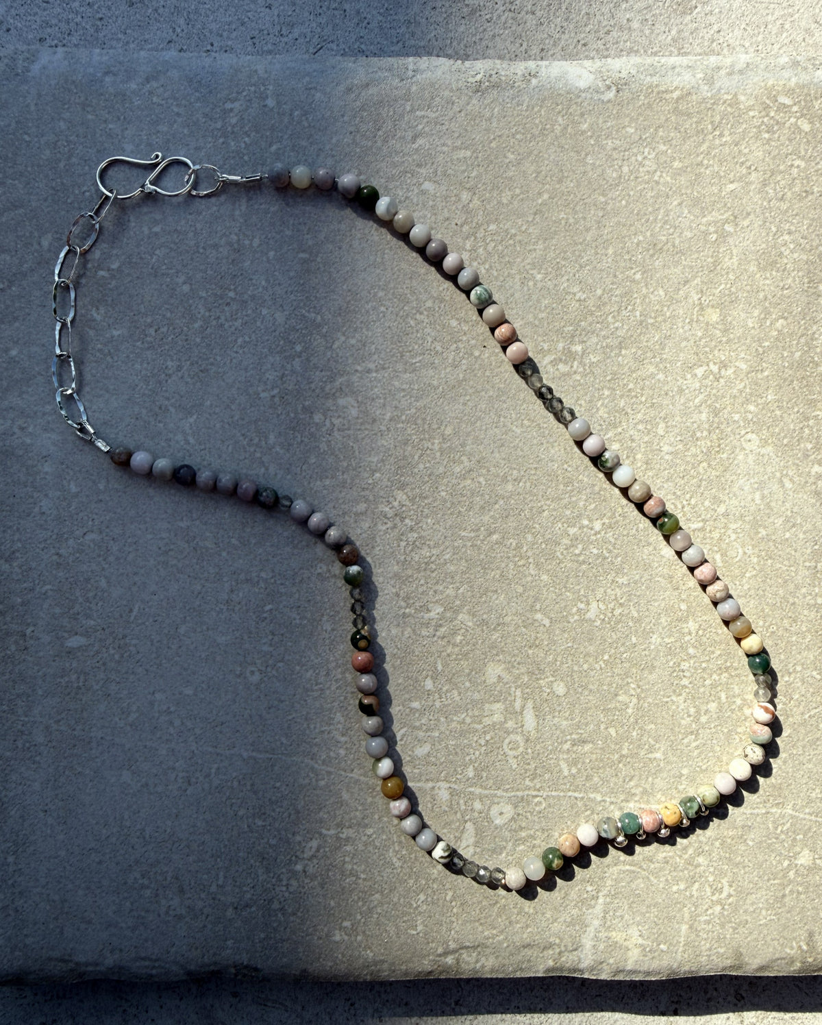 A beaded necklace made of small round beads in muted pink, gray, green, and brown tones, with a silver-toned chain extender and lobster clasp, laid on a textured stone surface.