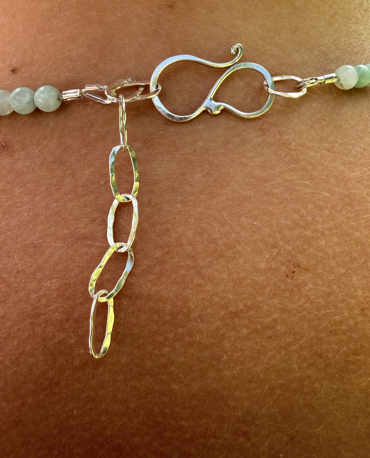 A close-up of a necklace worn on skin, showing a silver infinity-style clasp, a dangling hammered chain link charm, and light green faceted beads.