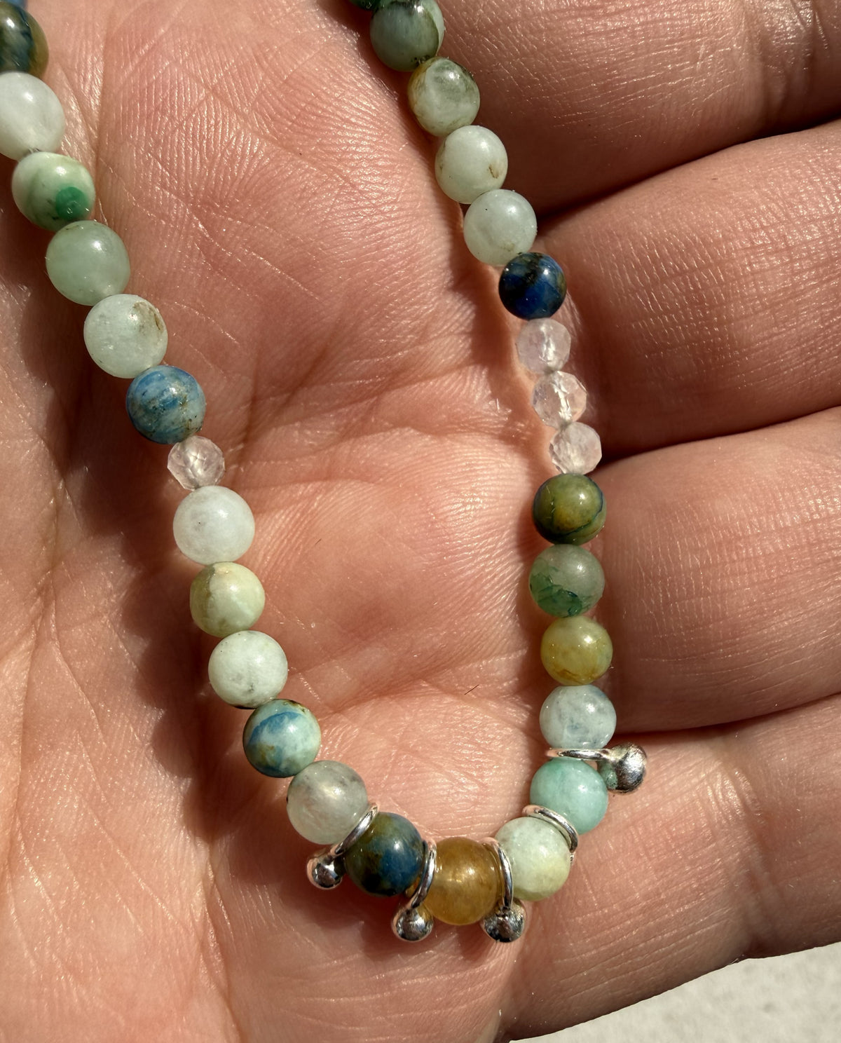 A beaded necklace made of polished round gemstone beads in shades of green, blue, white, and yellow, with small silver spacer beads, shown resting on a person's palm.