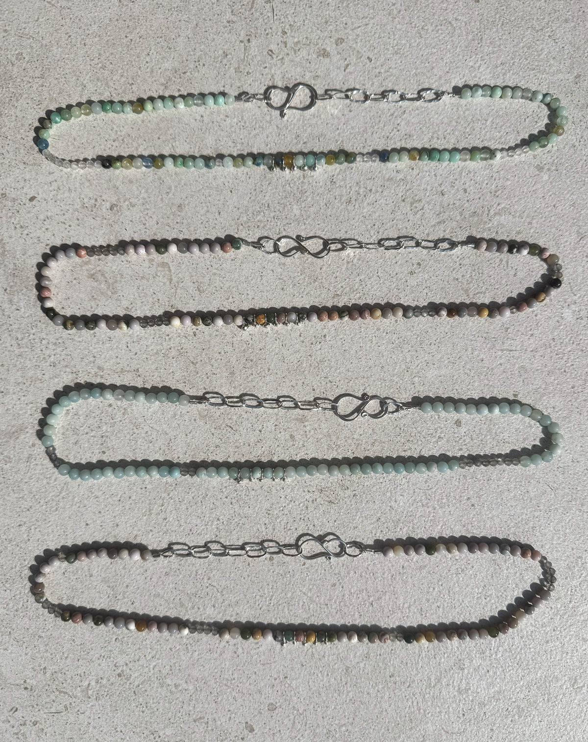 Four beaded necklaces laid flat on a light concrete surface, featuring small round beads in shades of gray, green, brown, and white, connected to silver-tone chain links with spring rings.