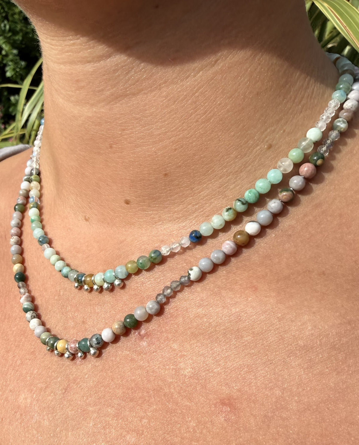 A close-up of a person's neck and chest wearing 2 beaded necklaces made of polished gemstone beads in pastel green, blue, pink, grey, and white.