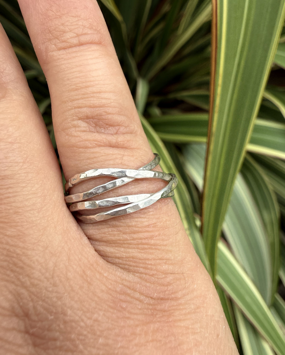 A close-up of a hand wearing four overlapping hammered silver rings, with green variegated plant leaves in the background.