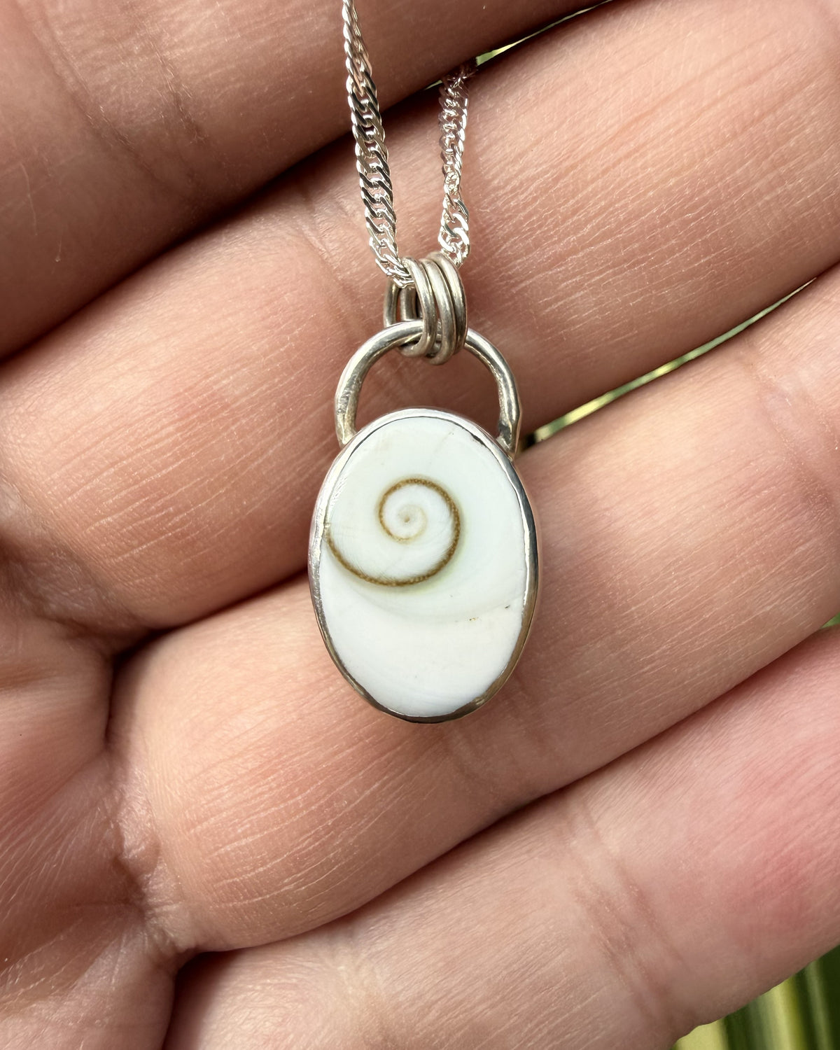 A silver oval pendant necklace featuring a white seashell with a brown spiral, held in a person's hand.