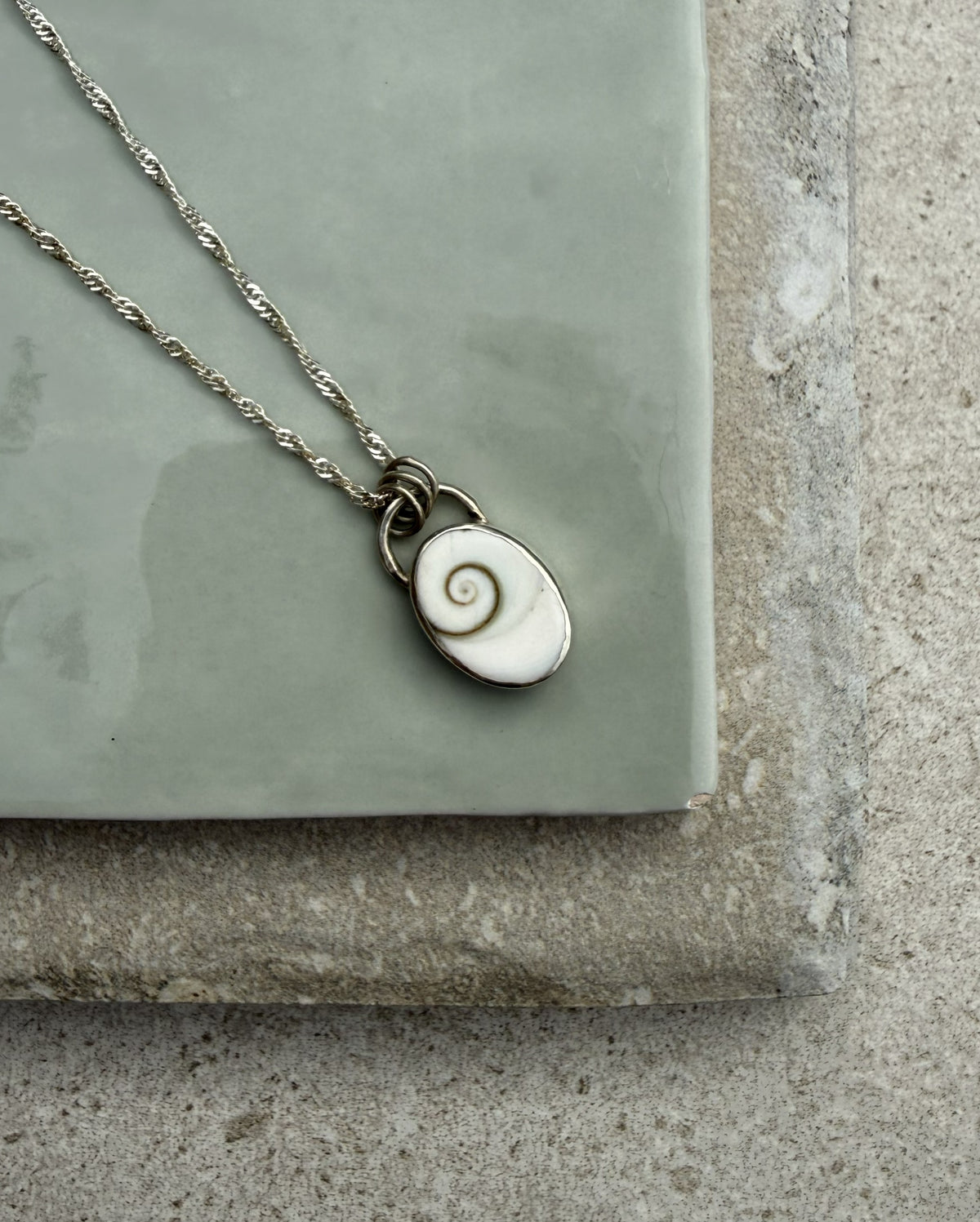 A silver chain necklace with a small oval pendant featuring a spiral shell design, displayed on a light green ceramic tile over a textured gray stone surface.