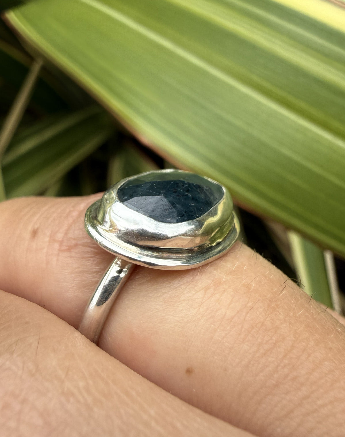 A polished sterling silver ring with a large, rounded, dome-shaped dark blue stone, shown worn on a person's finger with green leaves in the background.