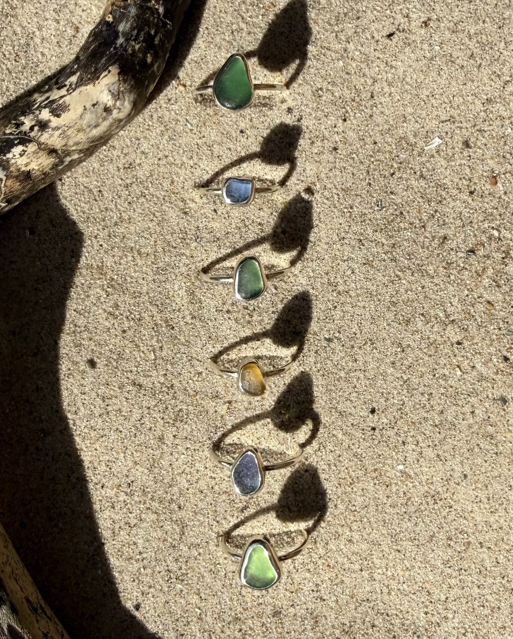 Seven sea glass rings in various colors are arranged in a vertical line on a sandy beach, casting long shadows on the sand.