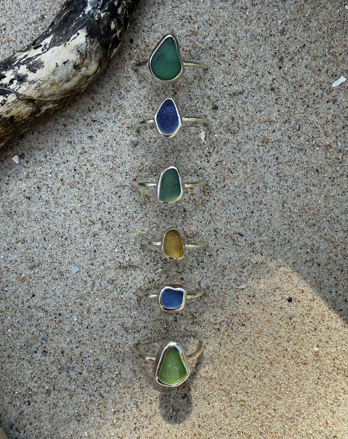 Six sea glass rings in various colors (green, blue, yellow) are arranged vertically on a sandy beach next to a piece of driftwood.