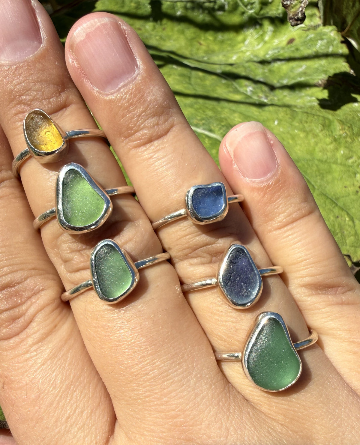 A close-up of a hand wearing six sea glass rings with teardrop-shaped stones in yellow, green, blue, and purple tones, set in silver, against a background of green leaves.