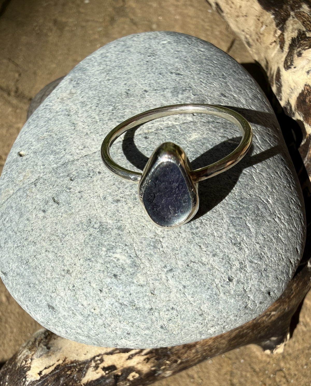 A silver ring with a polished, irregular dark gray stone, resting on a smooth gray river stone outdoors.