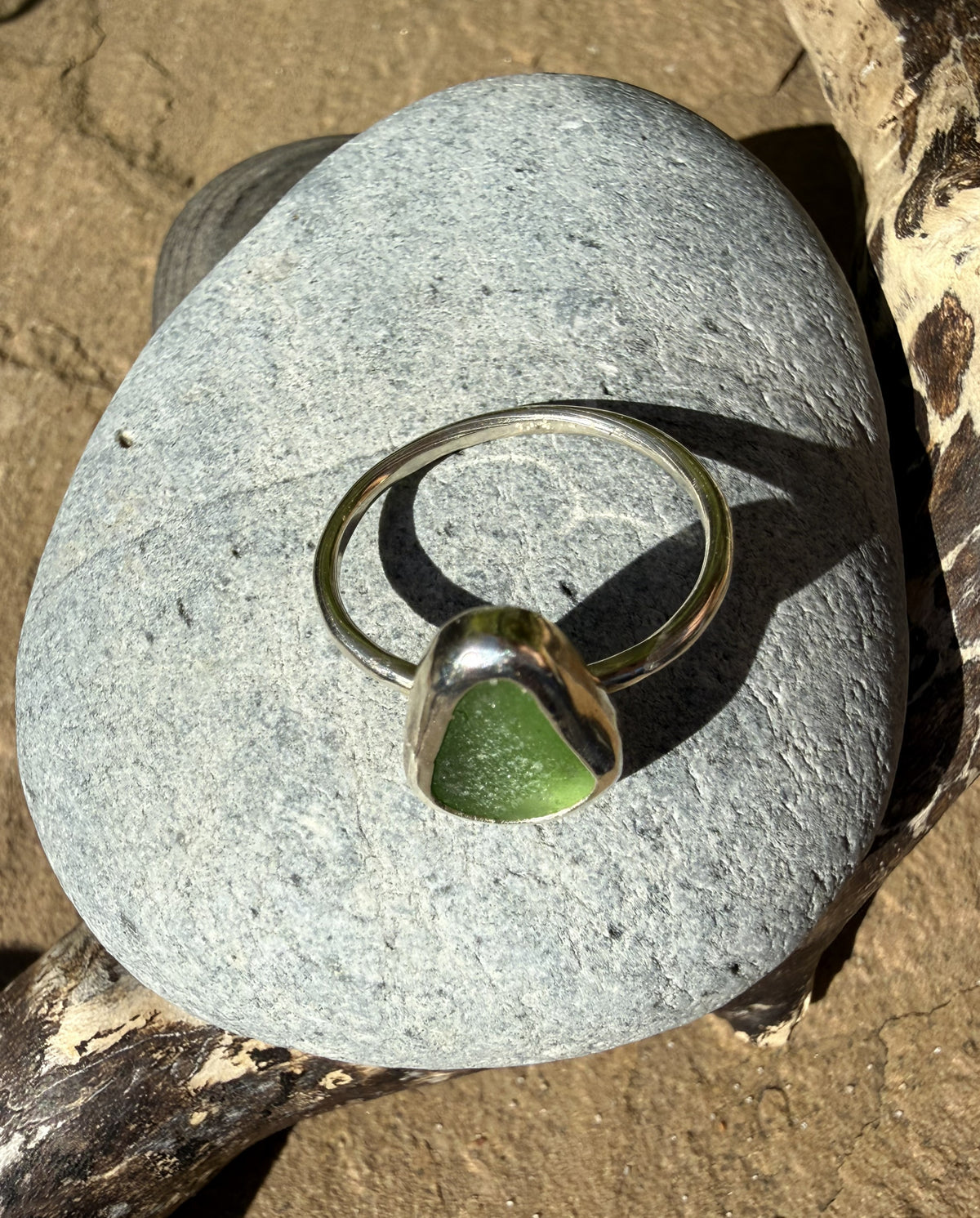 A polished silver ring with a smooth, organic-shaped green sea glass stone, displayed on a large gray stone outdoors.