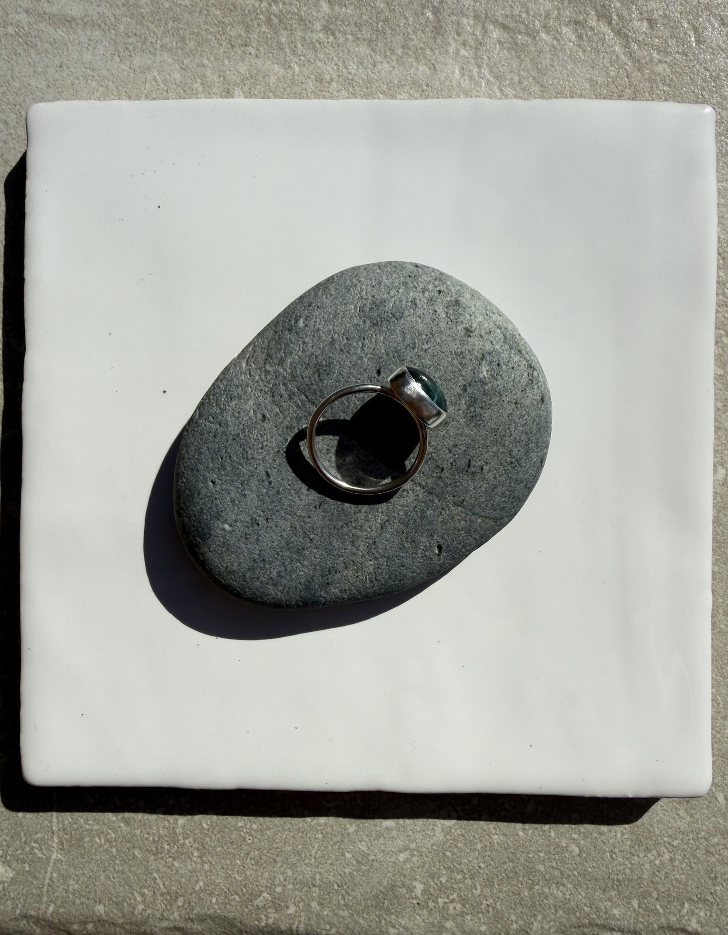 A silver-toned ring with a dark green stone is displayed on a smooth, dark gray stone, which is placed on a square white ceramic tile over a textured gray stone surface.