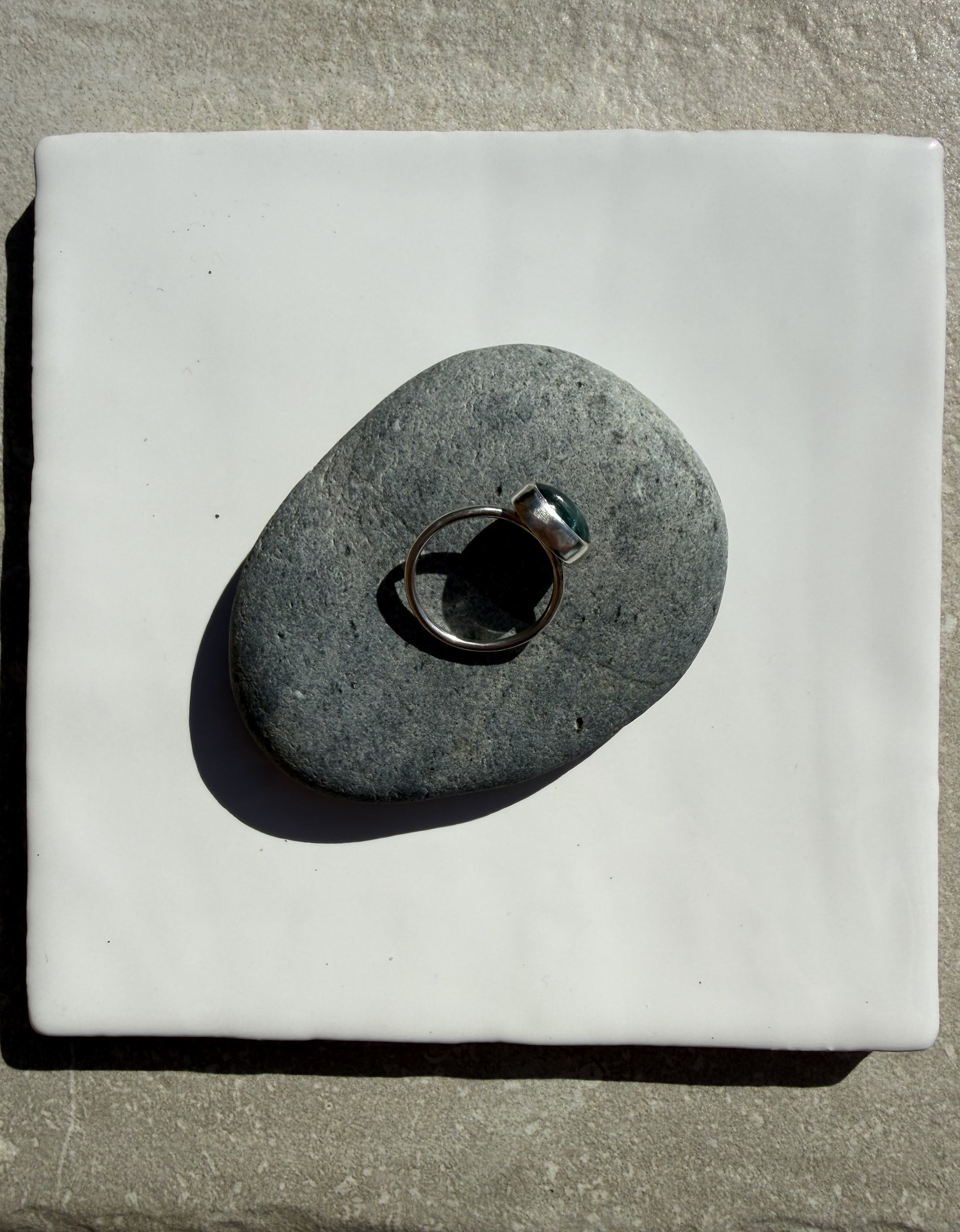 A silver-toned ring with a dark green stone is displayed on a smooth, dark gray stone, which is placed on a square white ceramic tile over a textured gray stone surface.